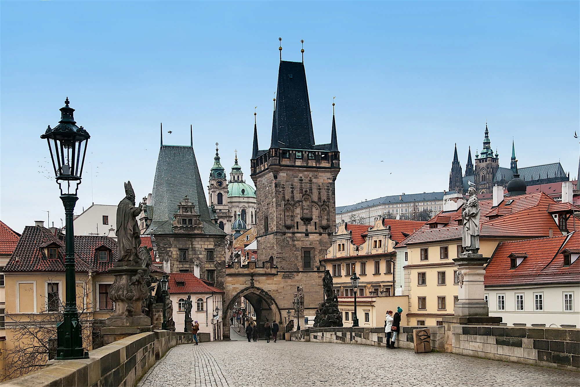 Historic stone bridge leading through a Gothic gate tower, lined with statues and red-roofed buildings under a clear blue sky.