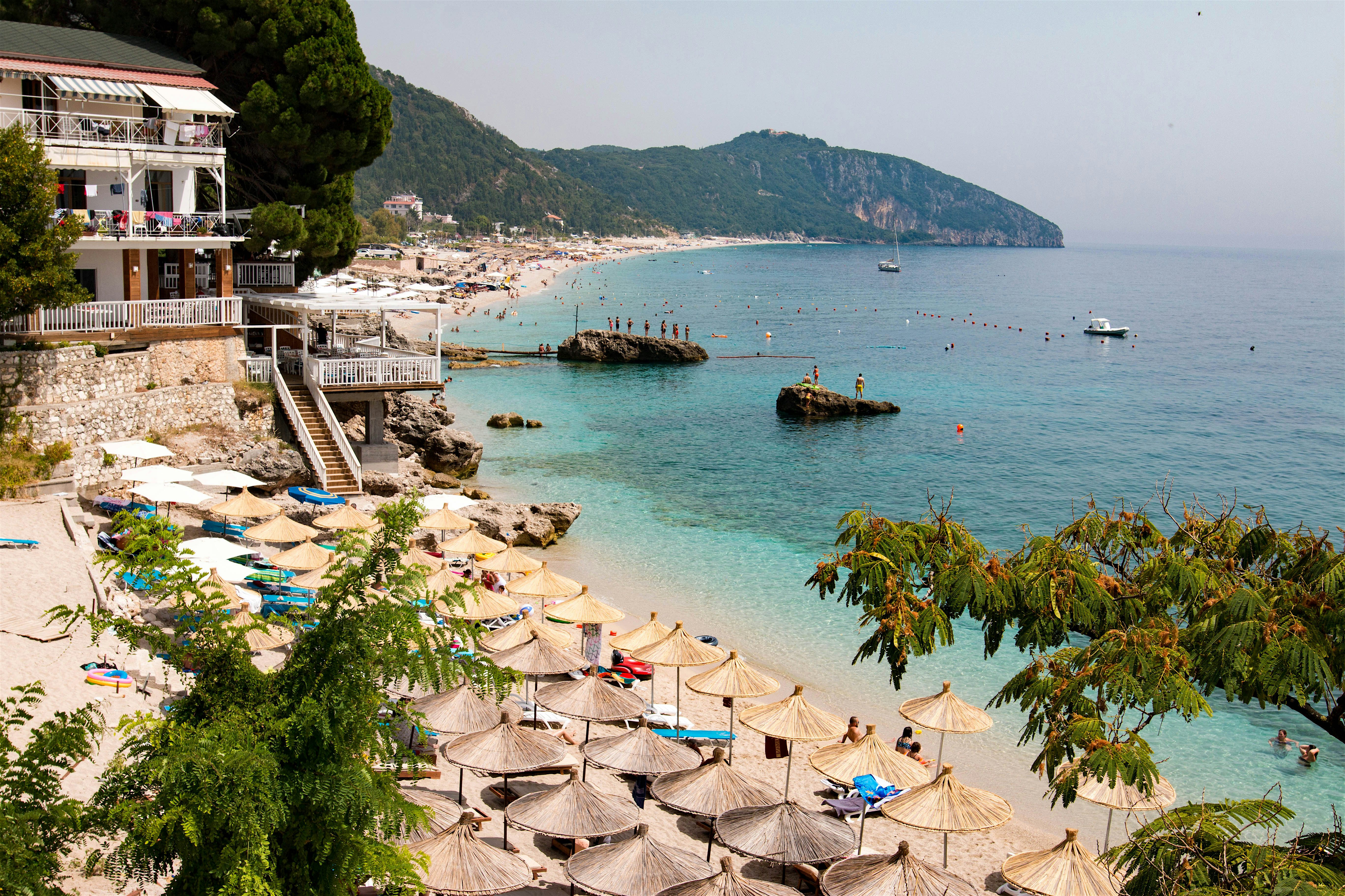 Dhërmiu Beach restaurant with steps down to straw parasols. Rocky coastline in the background curved around the sea.