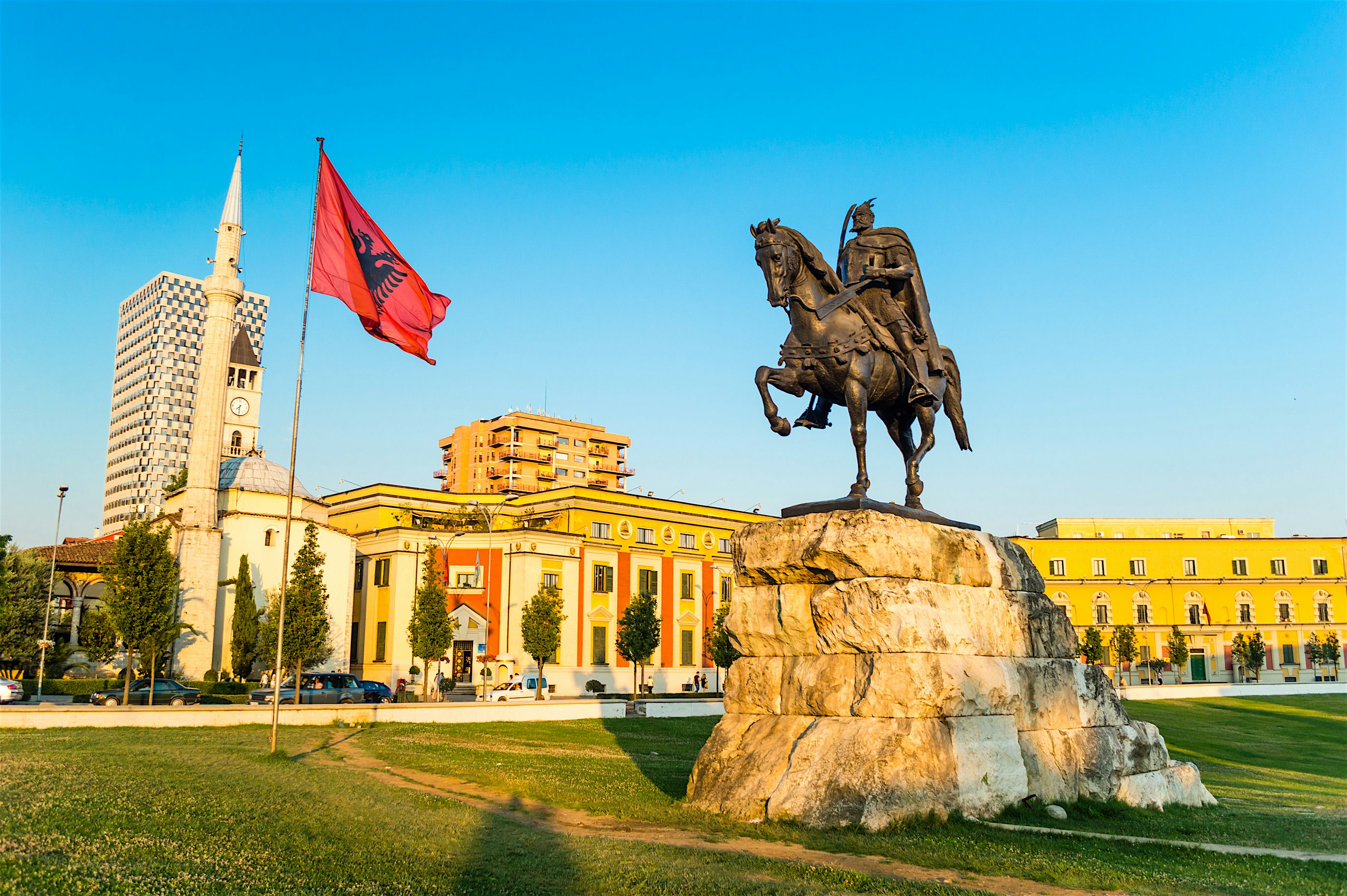 The square is lined with historic buildings, including the National History Museum, Skanderbeg Monument at the front which is a man on horseback, Et'hem Bey Mosque on the left and an Albanian Flag.