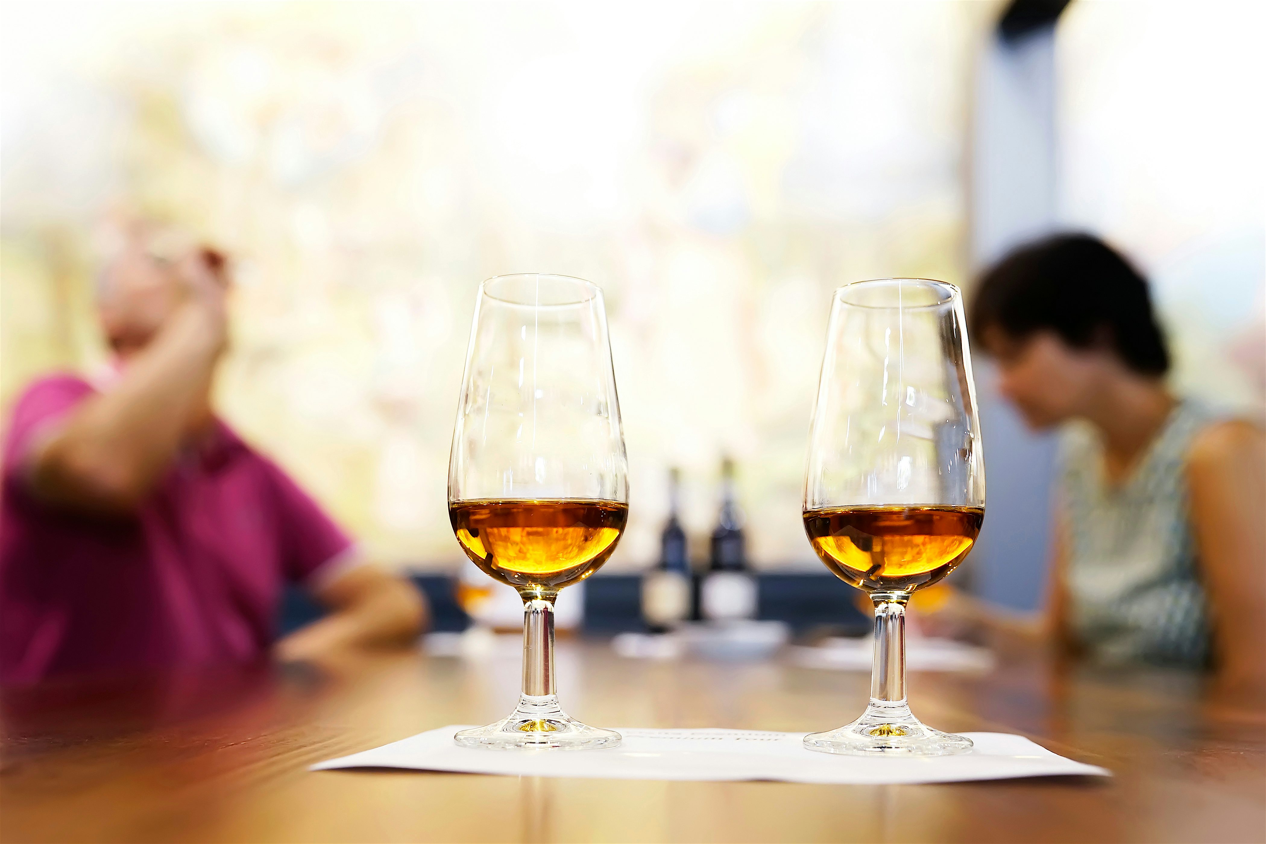 Two glasses at the front on a table as part of a Madeira wine tasting experience, to the rear are two blurred people with bottles at the far end