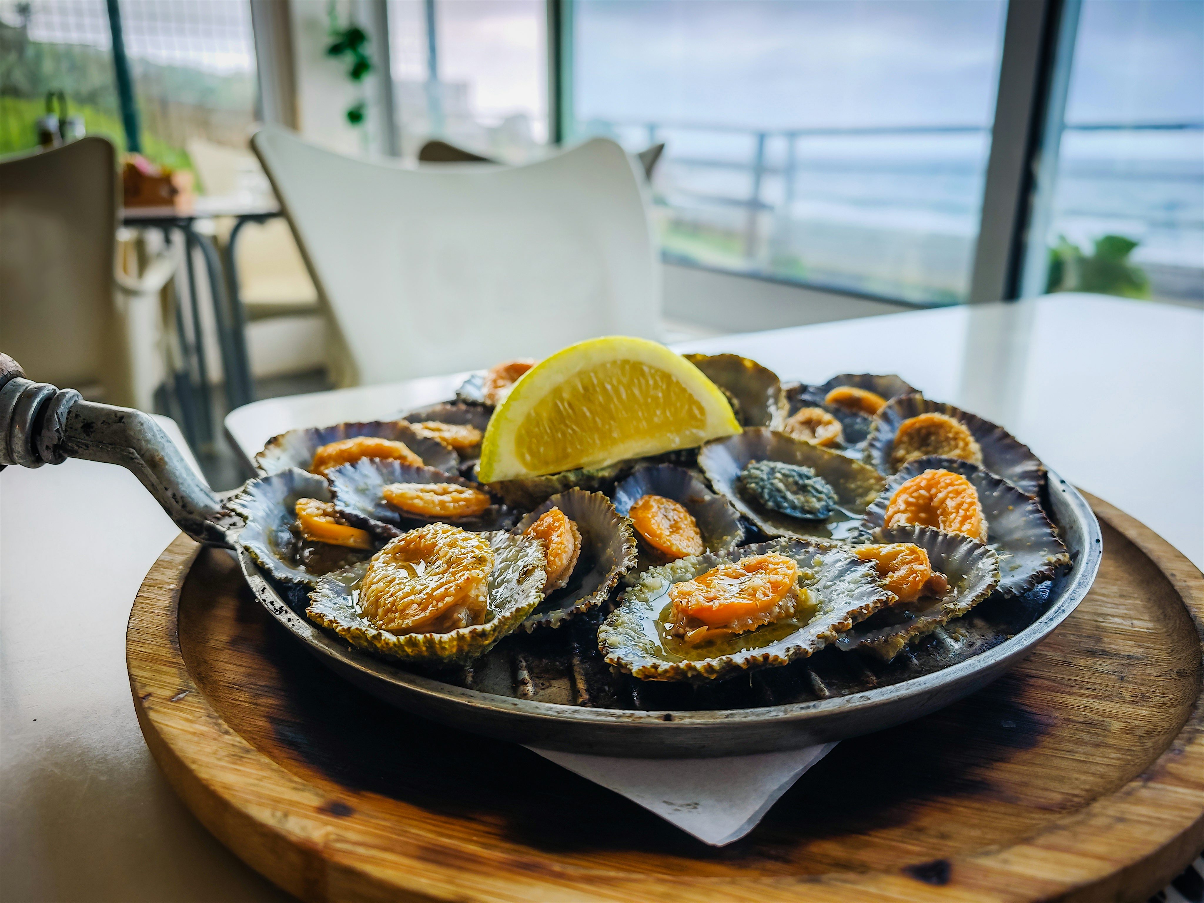 Cast-iron skillet with grilled limpets in their own shells with a lemon wedge on the top on a white table outside the restaurant is a sea view