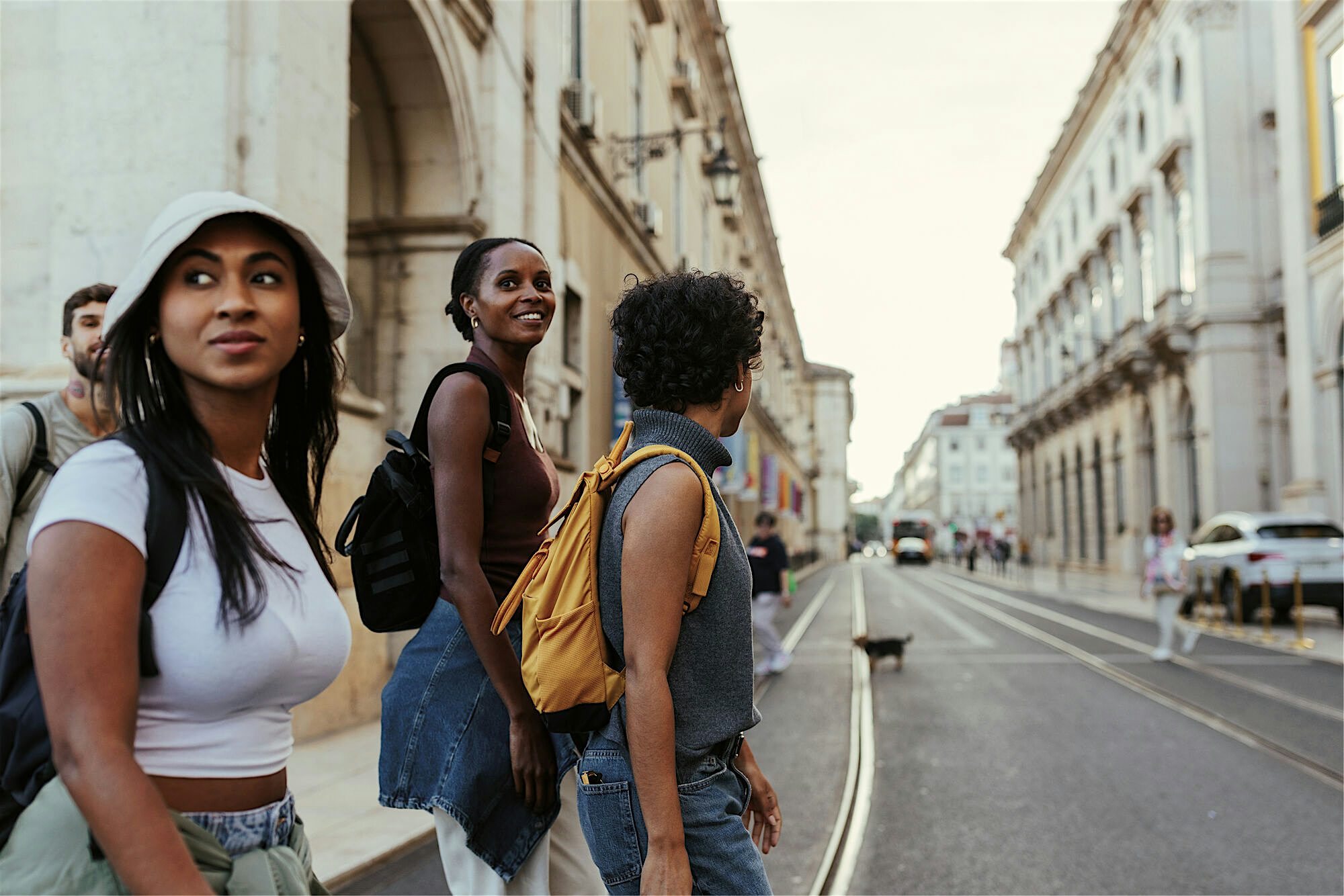 A small group walking along a wide city street lined with historic stone buildings, carrying backpacks and positioned near tram tracks.