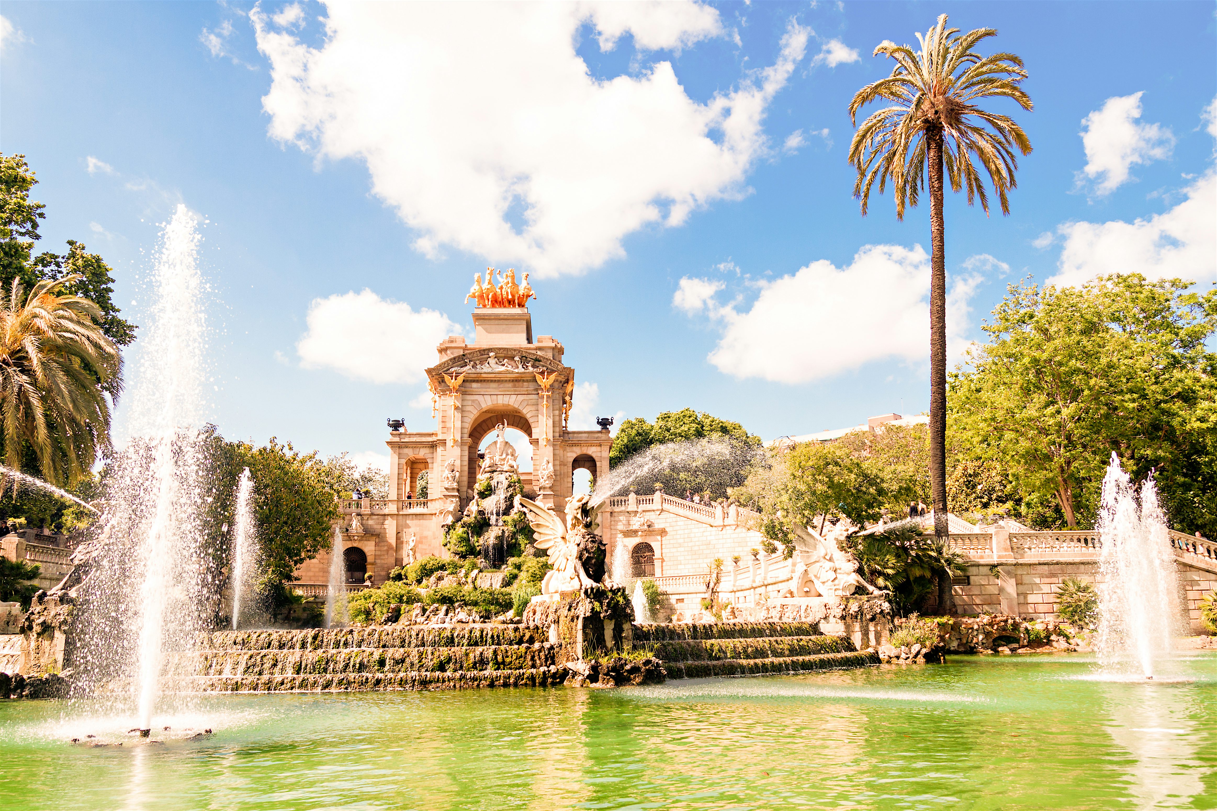 An ornate fountain and monument surrounded by palm trees and greenery, with water spraying into a large pool beneath a bright blue sky.