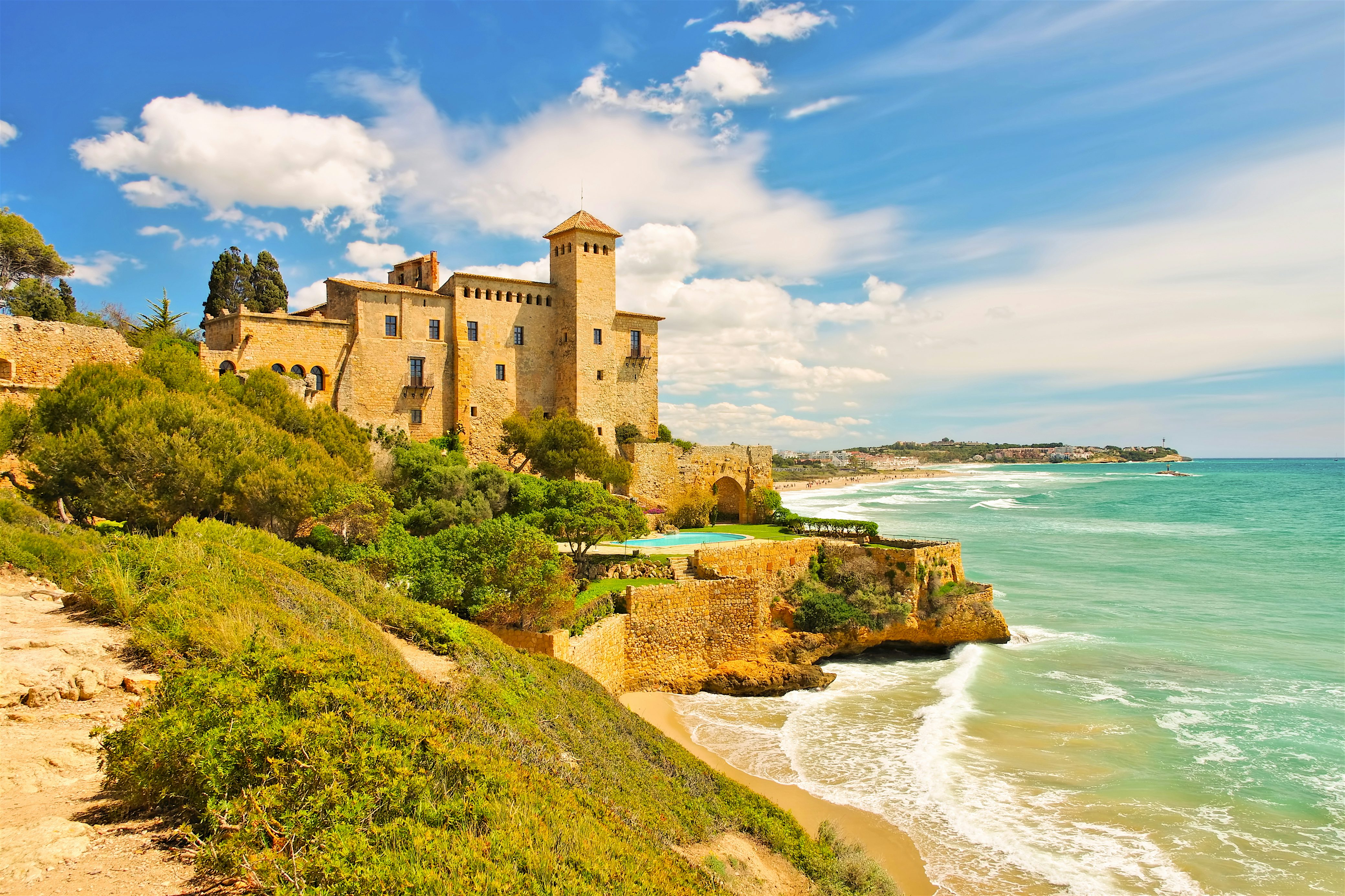 A historic stone building perched on a cliff above a sandy cove, with turquoise sea waves rolling in along the rocky coastline
