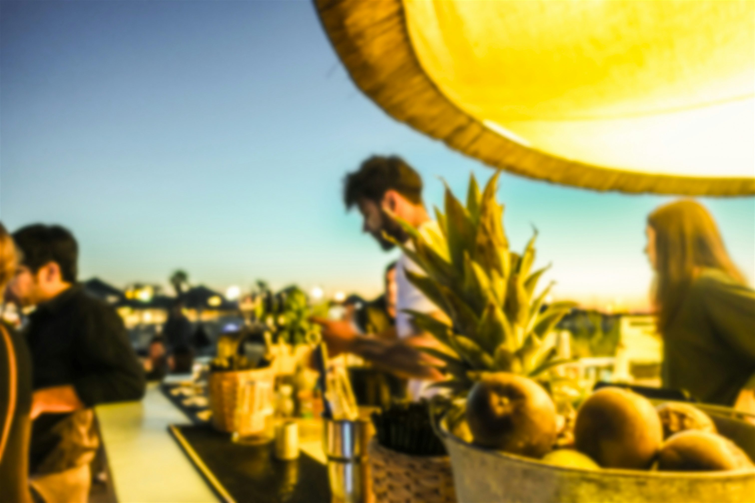 A rooftop bar setting with people preparing drinks beside a display of fresh fruit, overlooking a city at sunset under warm lighting.