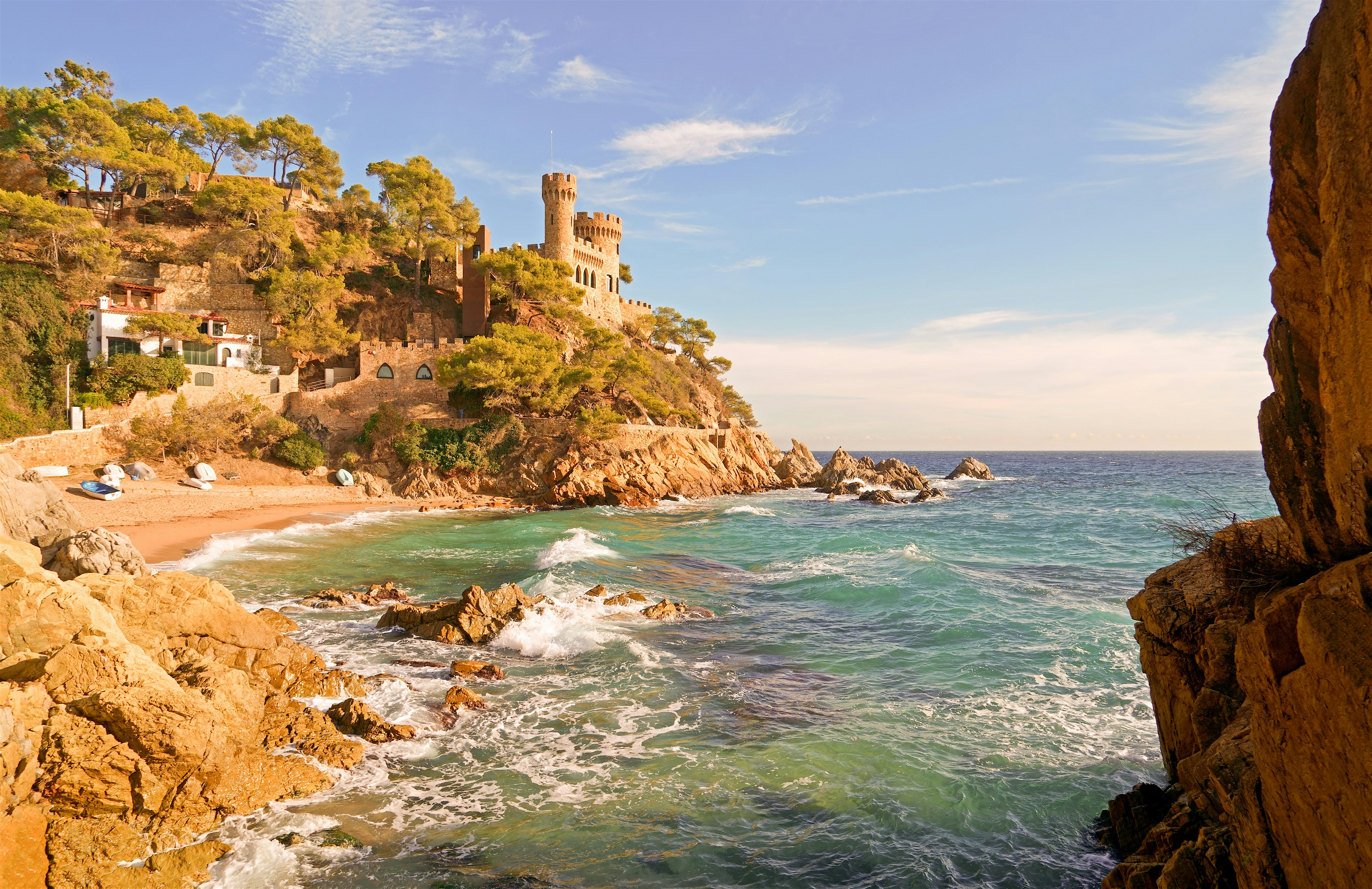 A small beach framed by rocky cliffs and pine trees, with a castle overlooking the shoreline and calm blue water stretching out to sea.