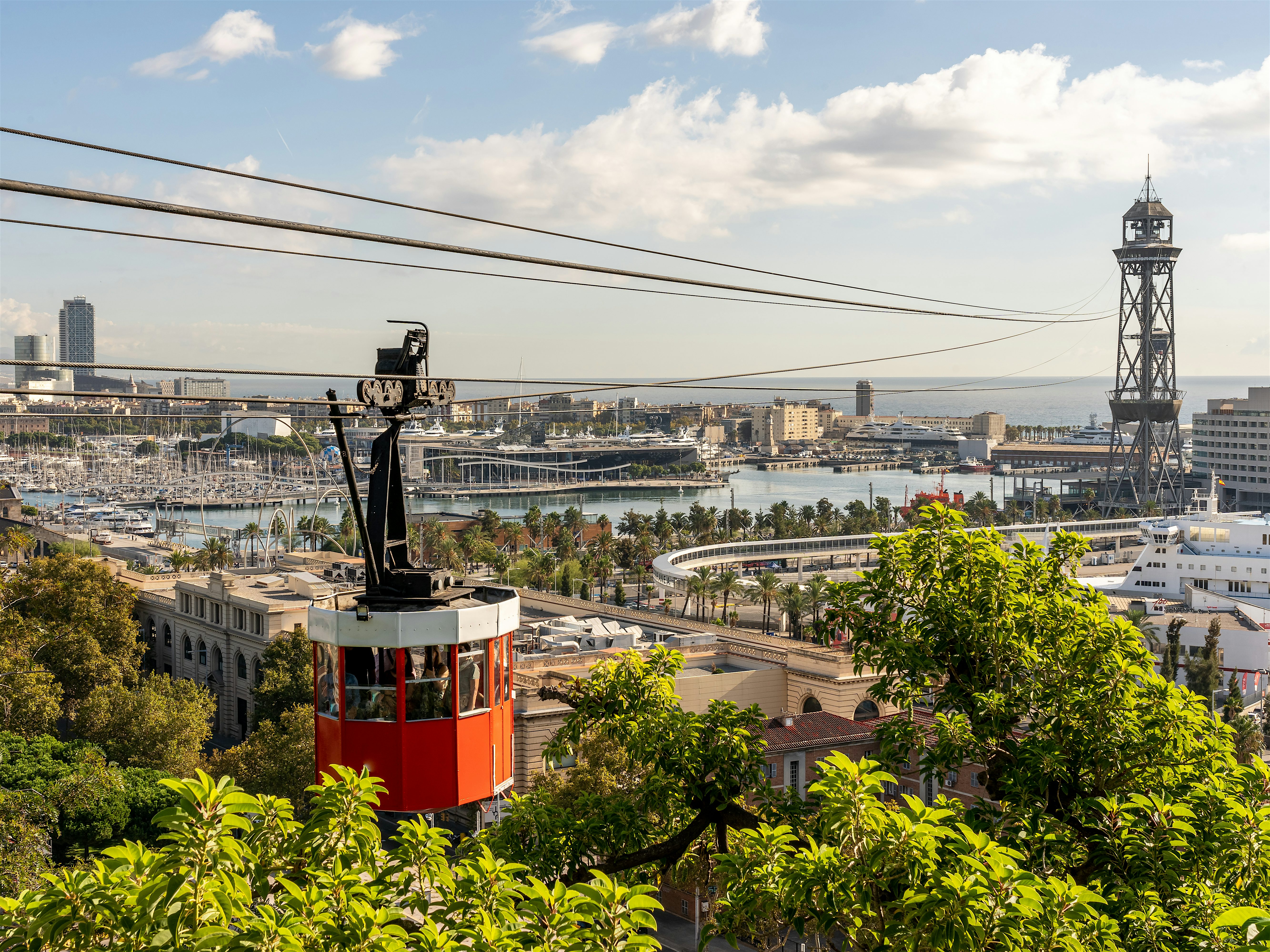 A red cable car suspended over a lush hillside, overlooking a coastal city with a marina, cruise ships, and a tall metal tower near the waterfront. In the background, the sea and a partly cloudy sky frame the urban skyline.