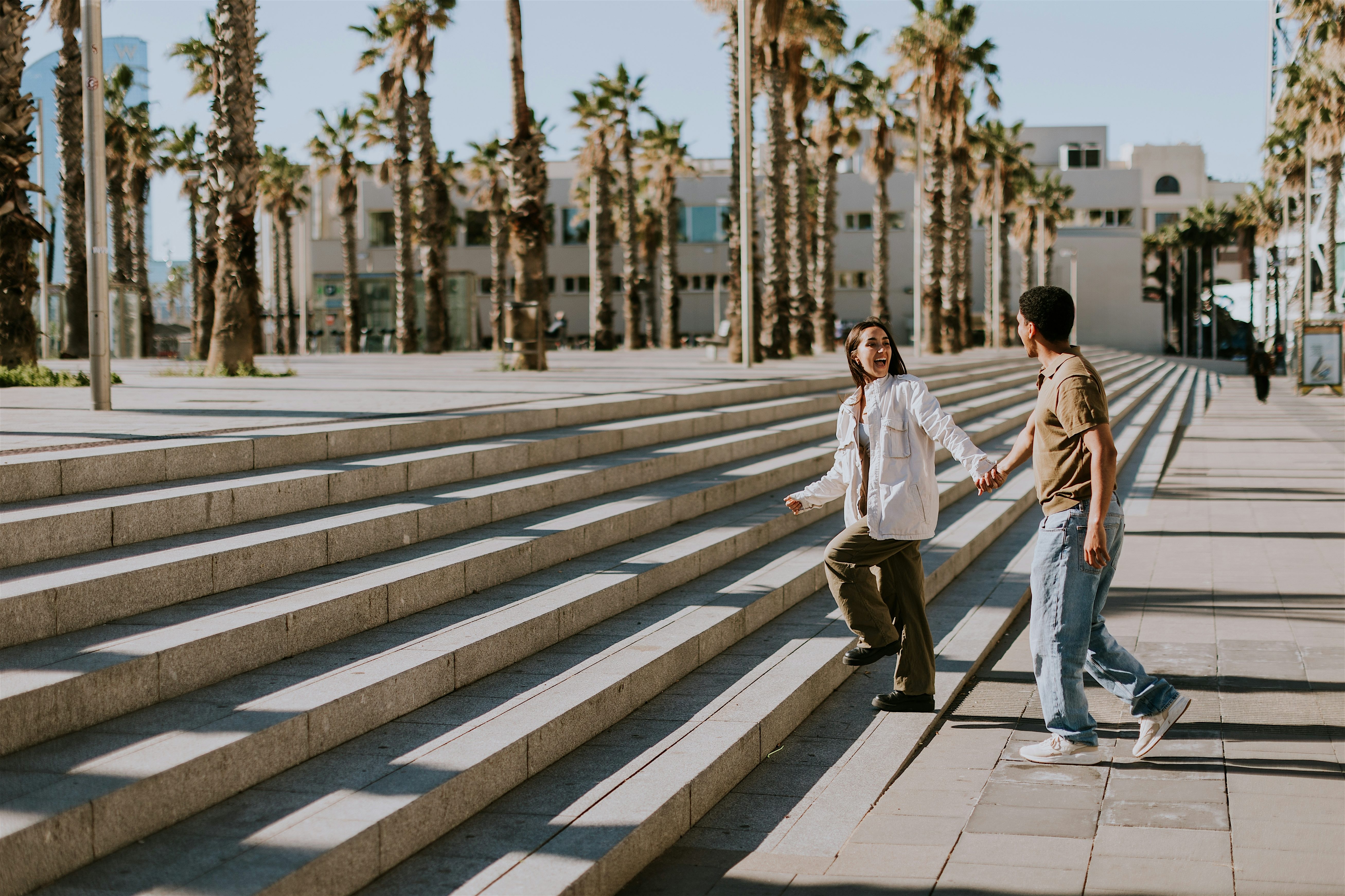 A couple hand in hand walking up an outdoors staircase, the street is line with palm trees, but empty aside from that. The day is bright and a clear blue.