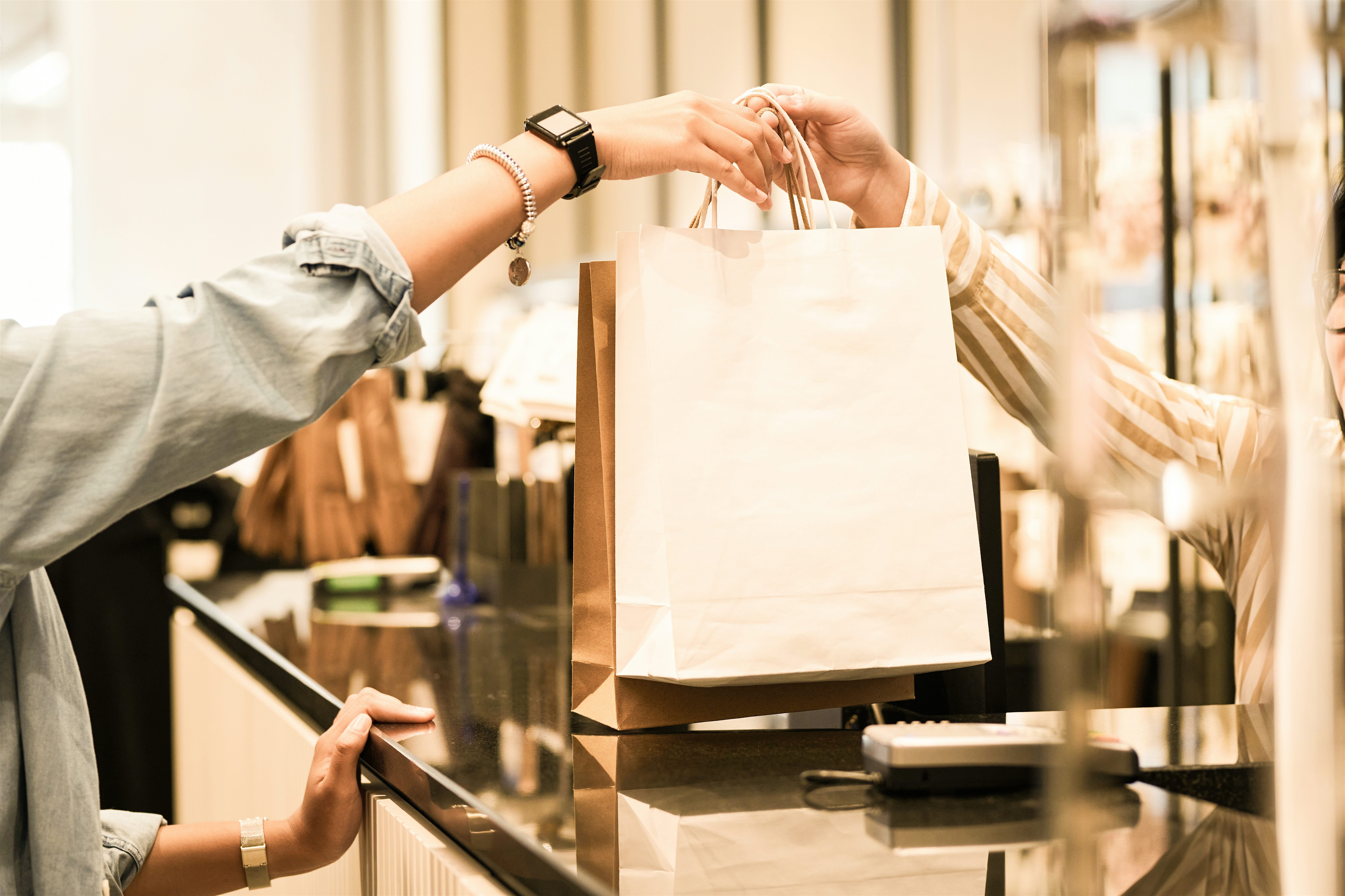 A paper shopping bag being handed over a retail counter, with hands reaching across the till in a modern shop.