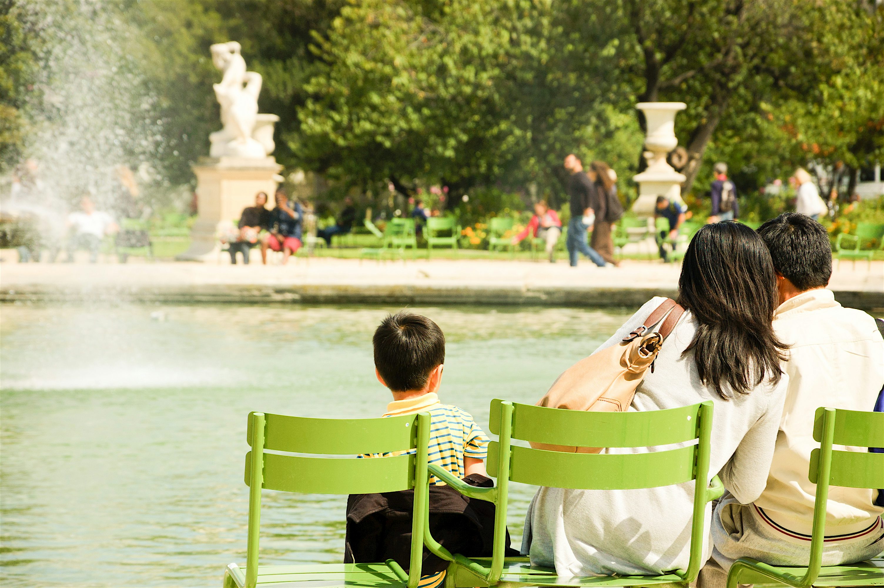 People sitting on green chairs beside a fountain in a city park, watching the water with statues and trees in the background.