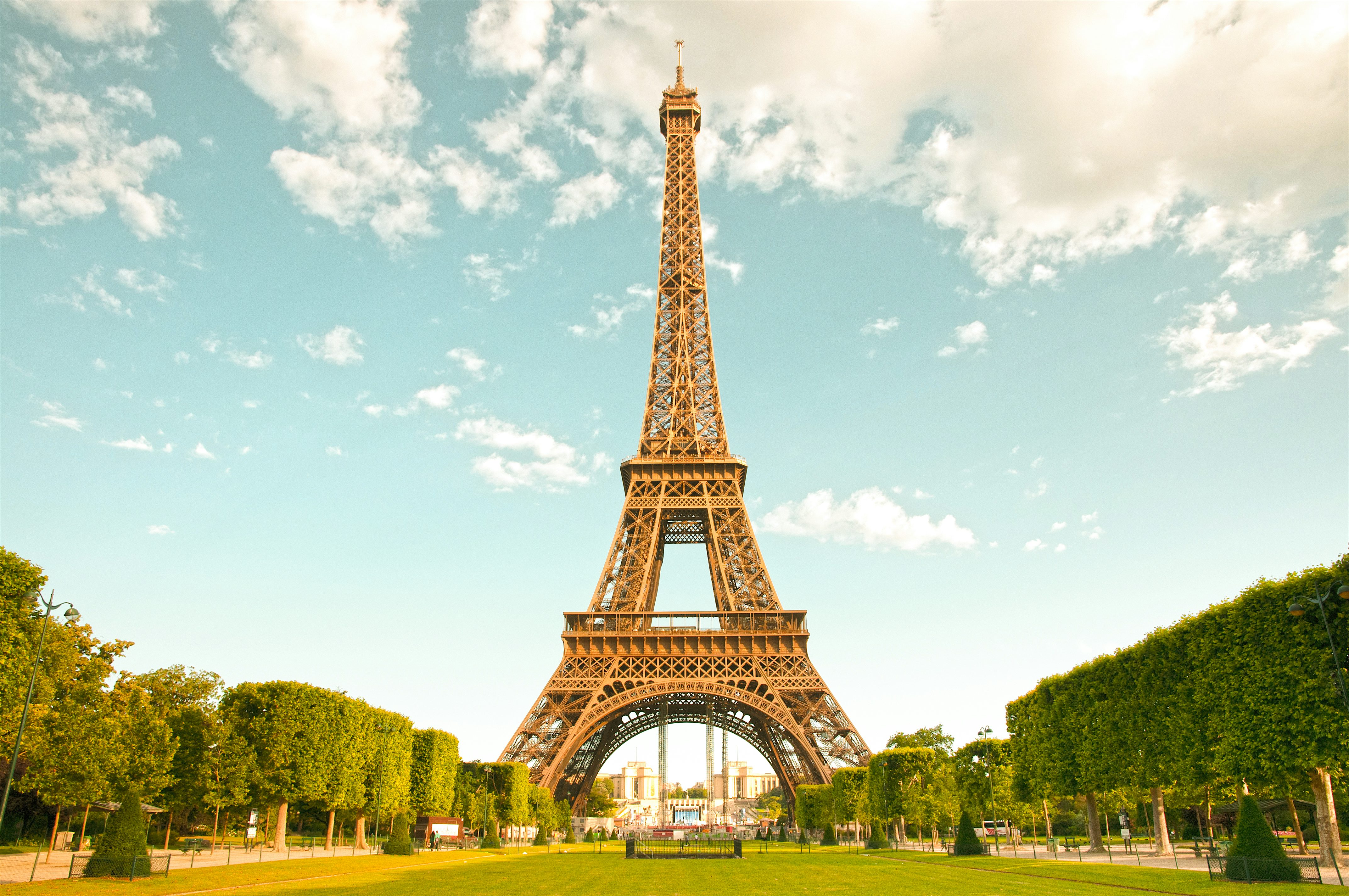The Eiffel Tower rising above a manicured park, framed by rows of green trees under a clear blue sky.