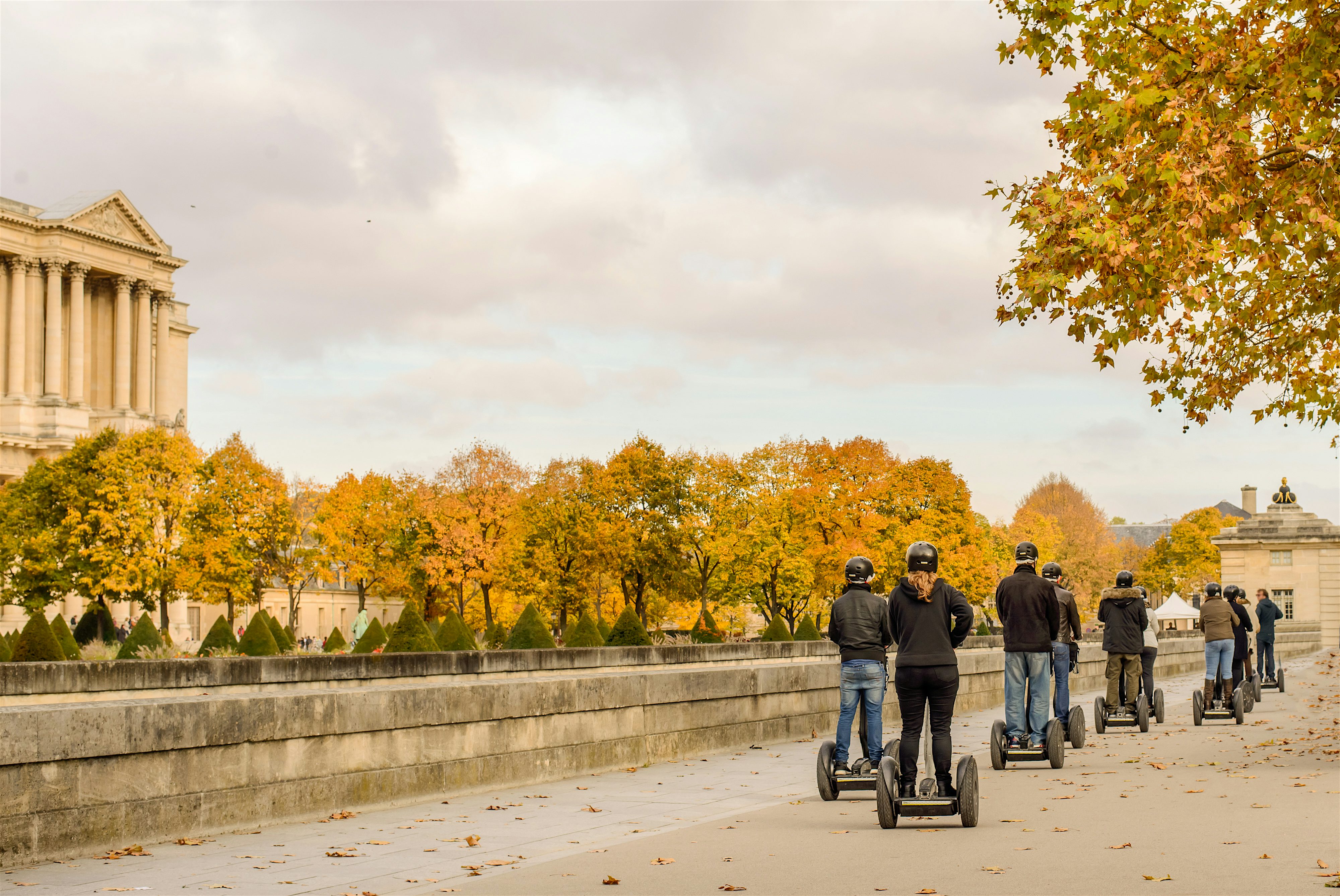 Group of people riding Segways along a wide stone path, lined with autumn trees and historic buildings in soft afternoon light.