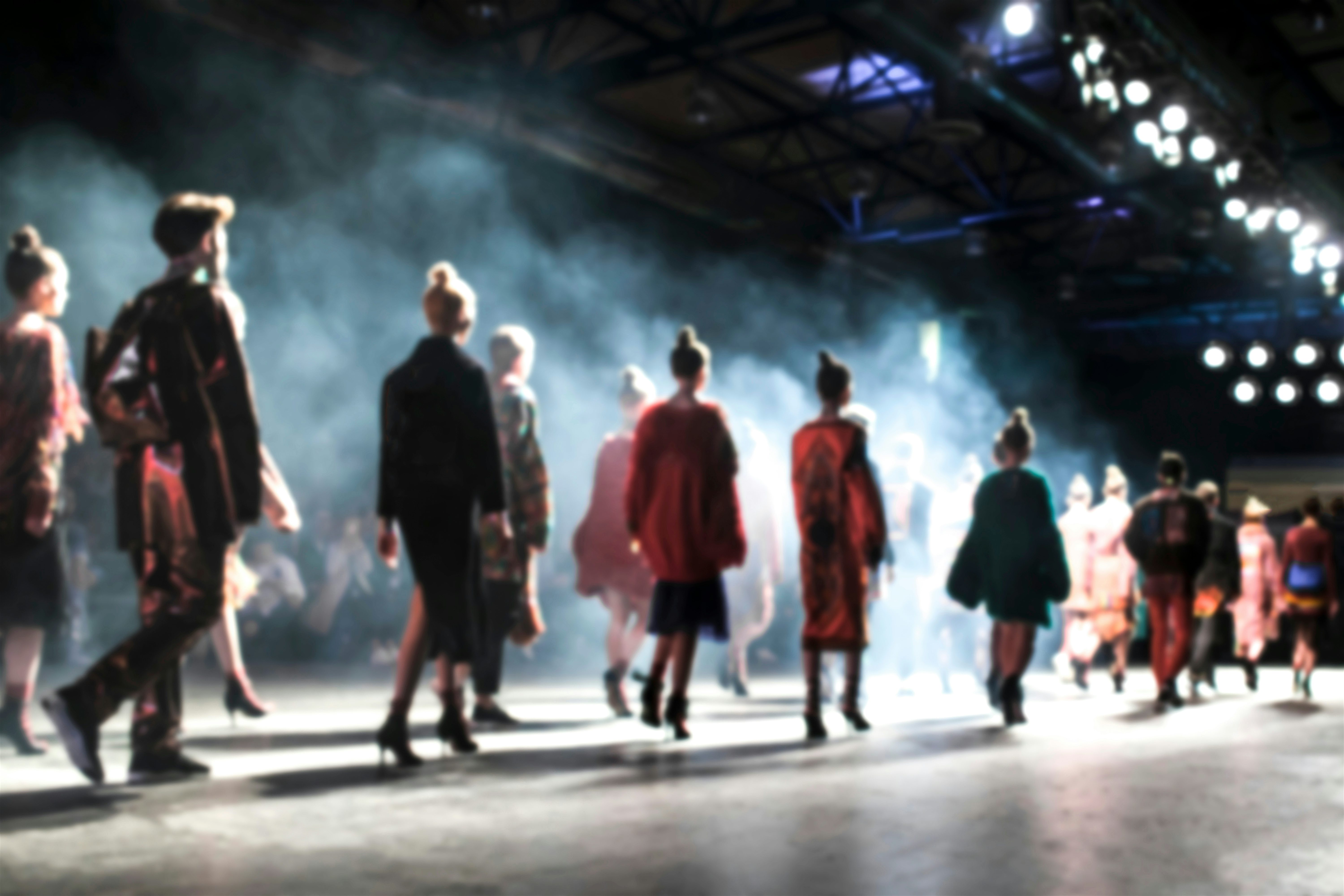 Models walking down a runway during a fashion show, stage lights and light haze creating a dramatic atmosphere.