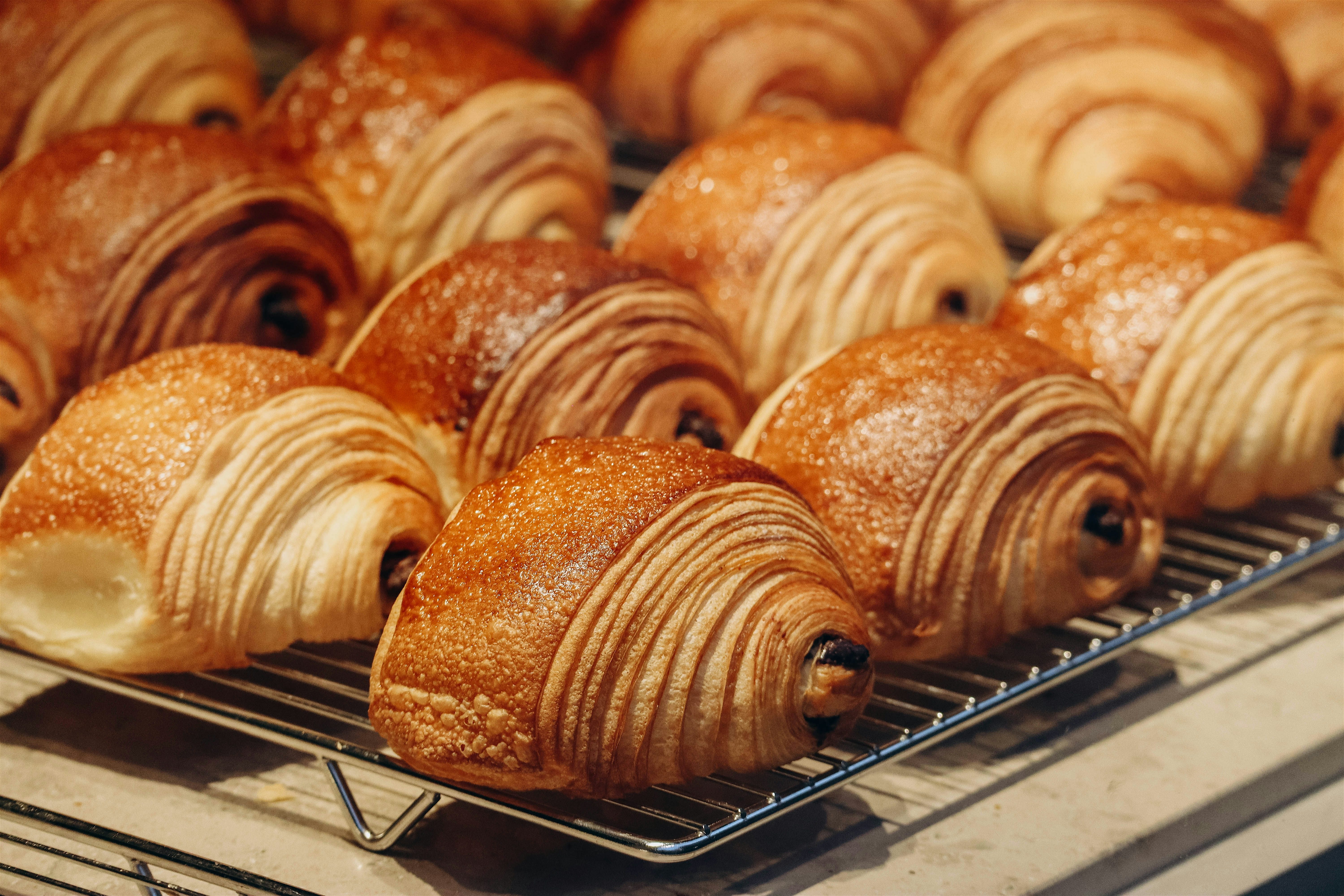 Rows of golden croissants cooling on metal racks, their flaky layers and crisp edges catching the light in a bakery display.