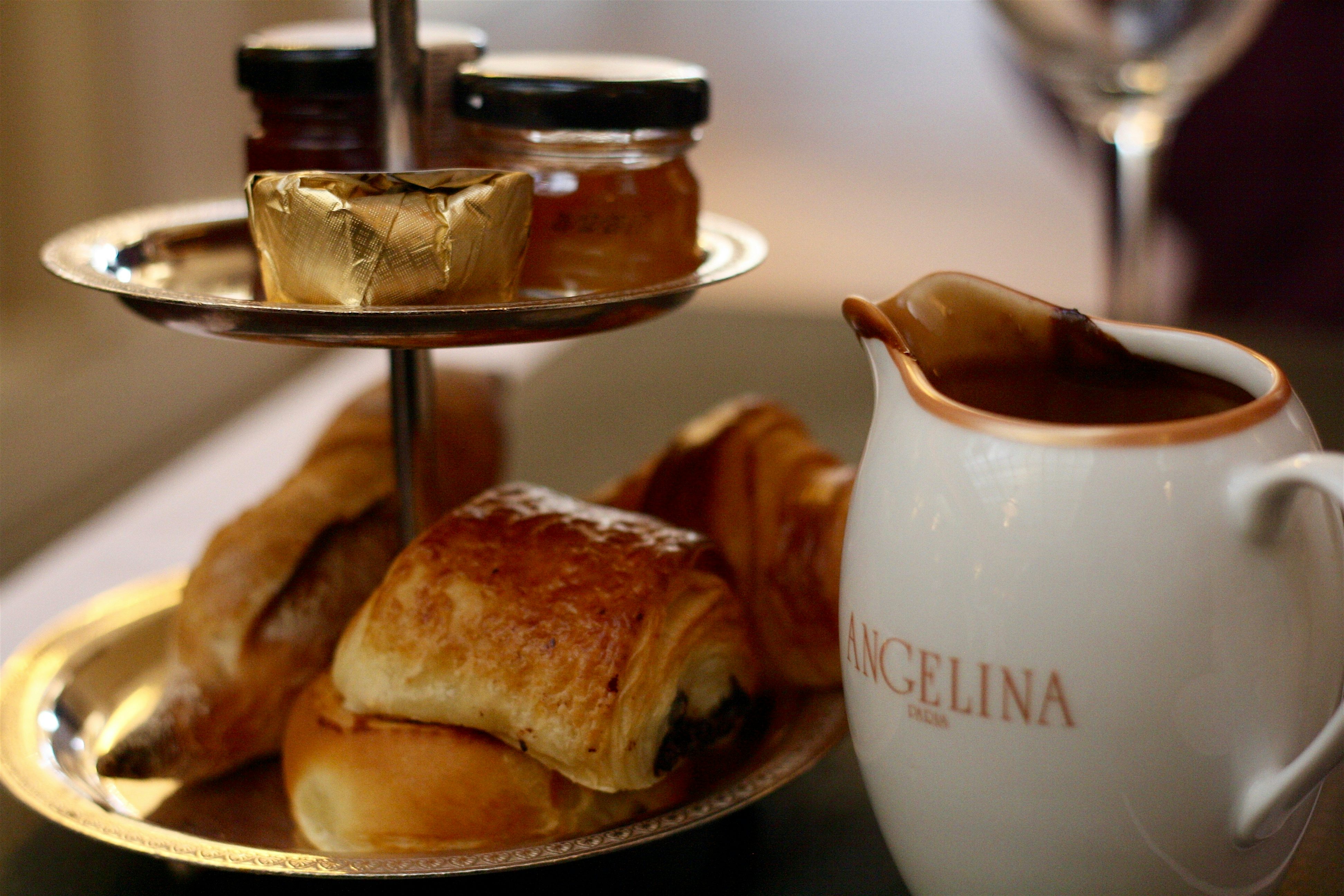 Pastries and jars of jam served on a tiered stand beside a ceramic jug of hot chocolate, arranged on a café table.