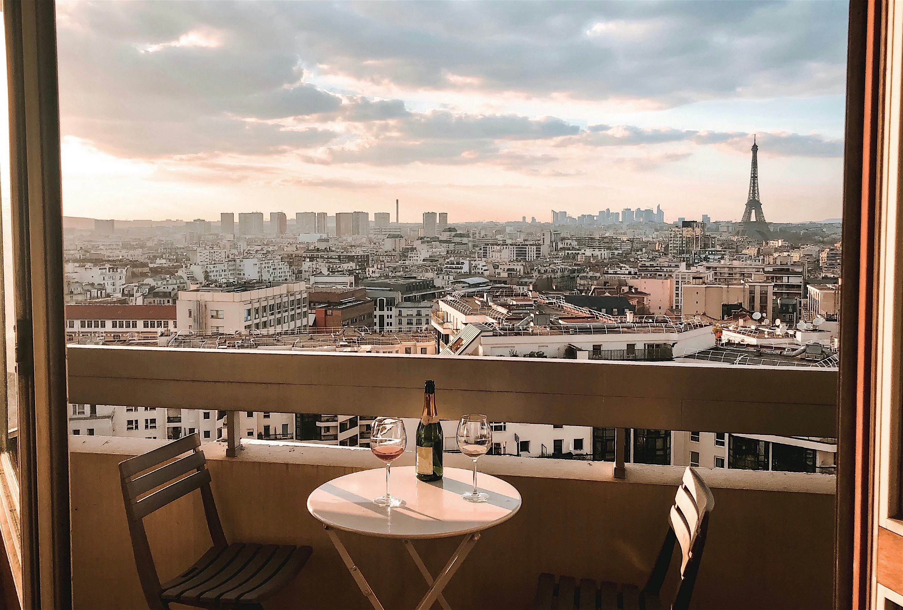 Balcony view over Paris at sunset, with a small table set with wine glasses and the Eiffel Tower visible on the horizon.