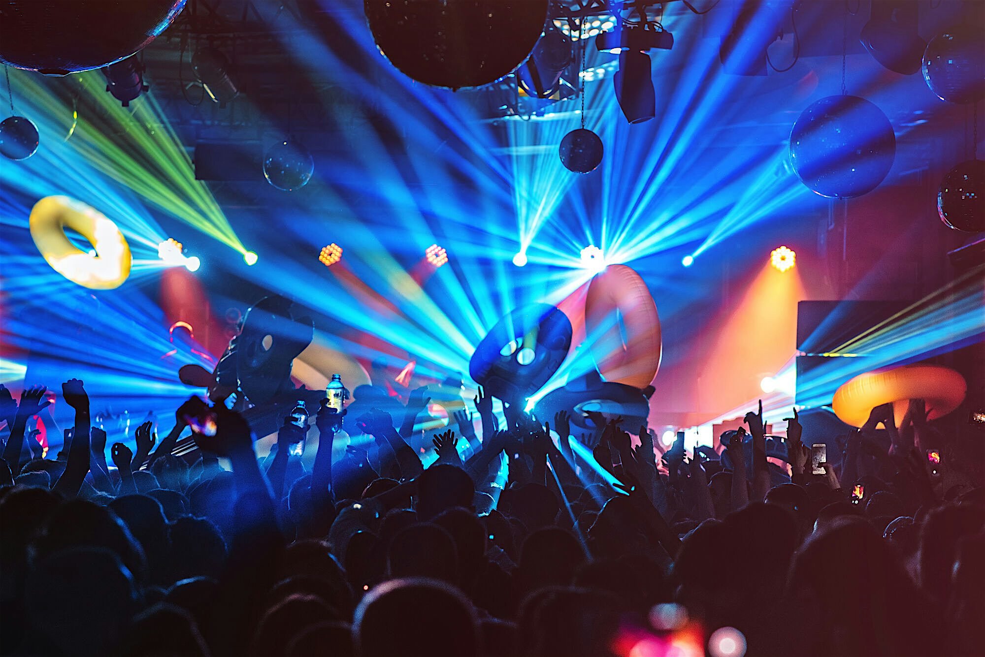 Crowded nightclub scene with hands raised towards a stage, bright laser lights and colourful inflatables filling the space above the audience.