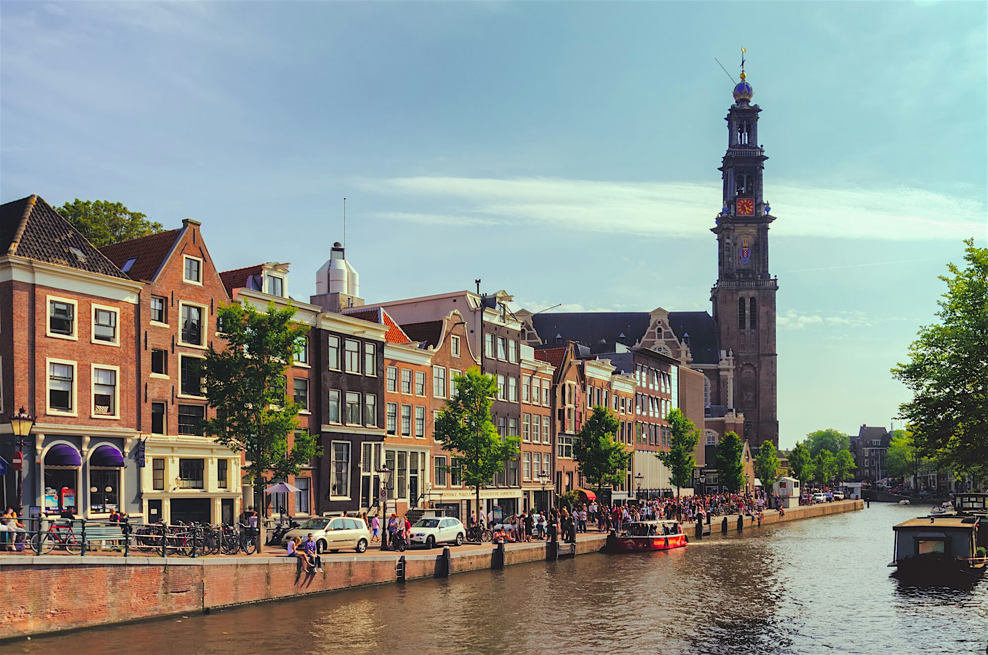 Canal lined with traditional buildings and trees, with people walking and sitting along the waterfront and a tall church tower