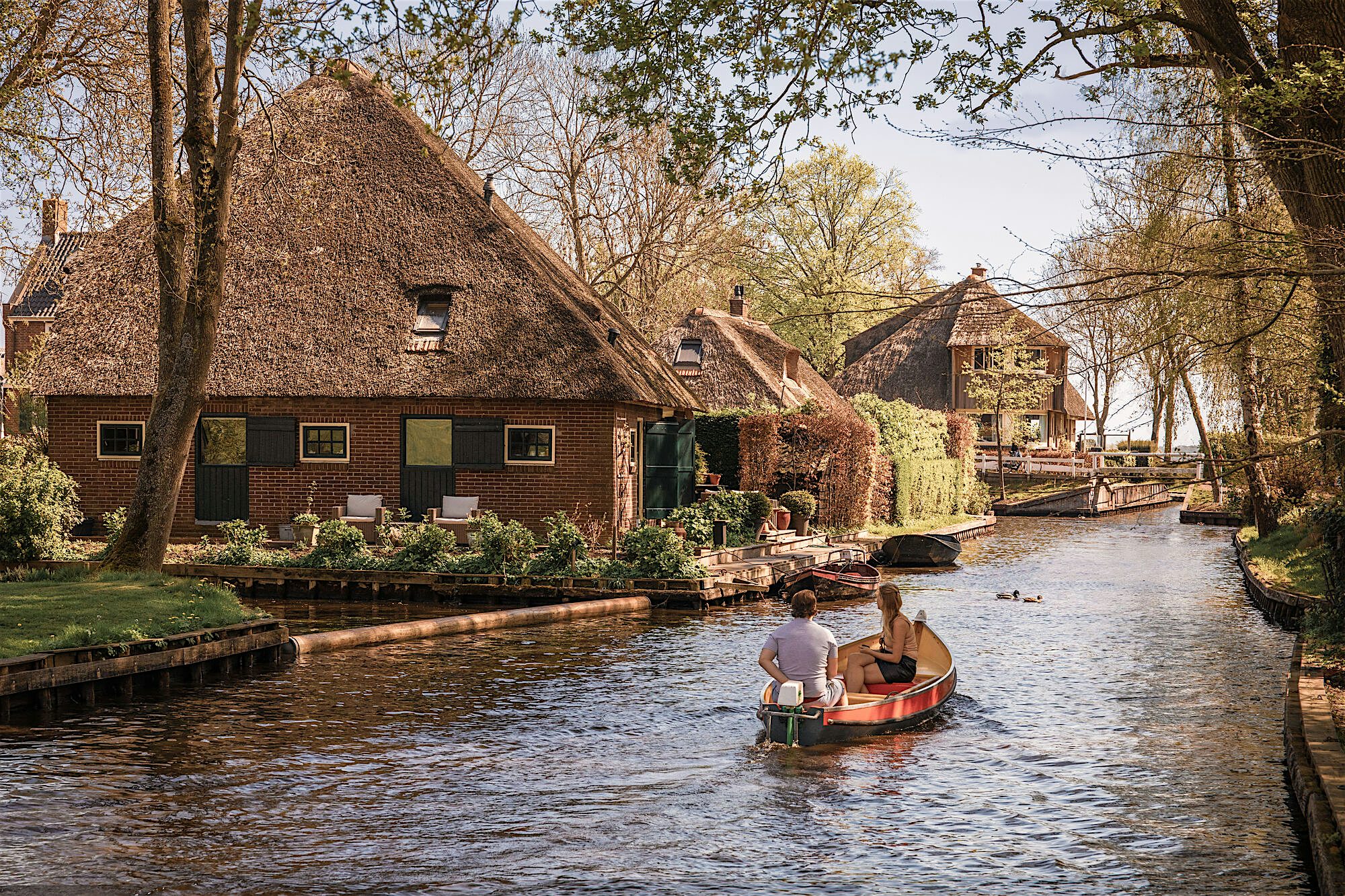 Small boat carrying two people along a narrow canal bordered by traditional thatched cottages and trees,