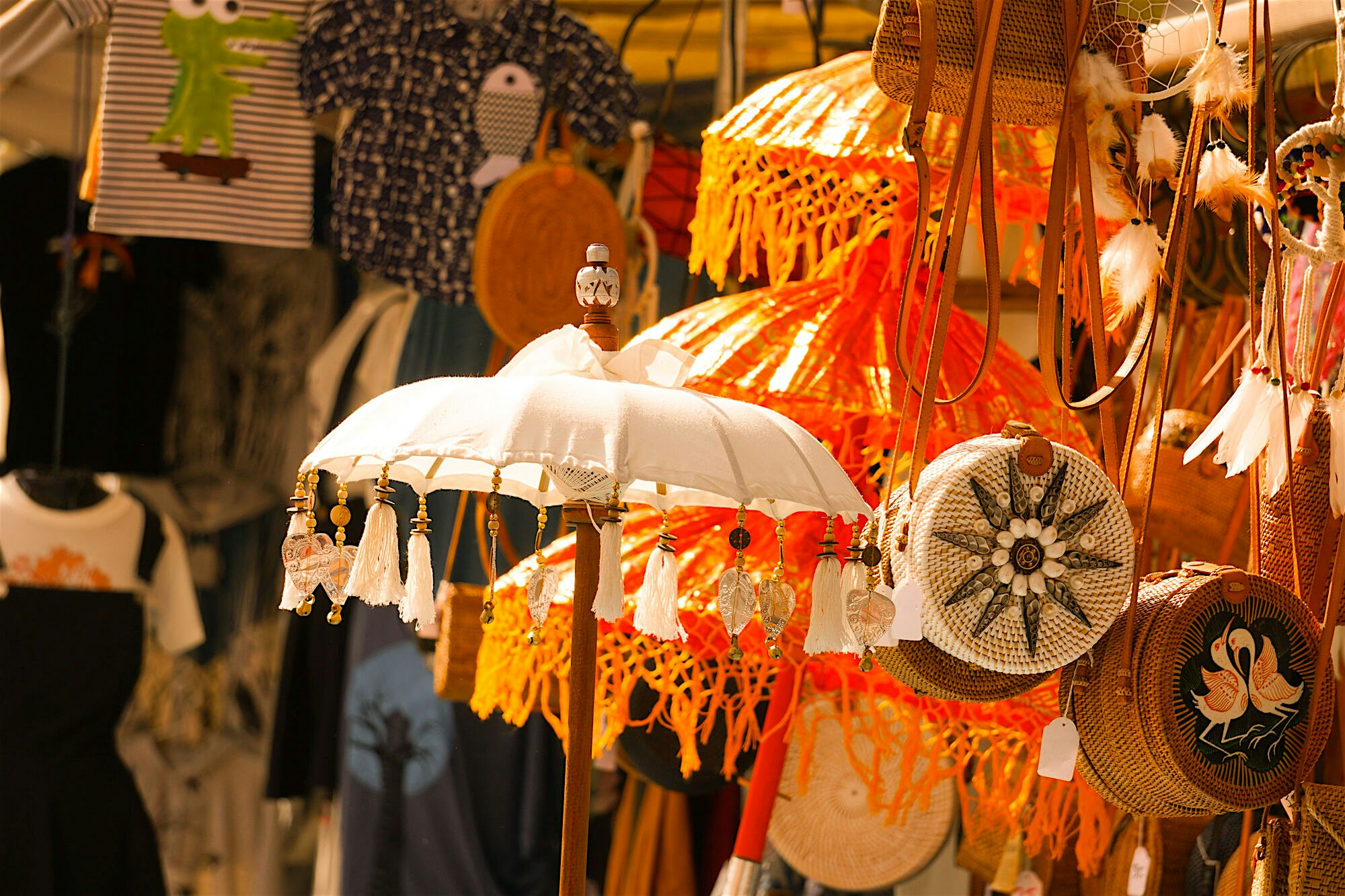 Market stall displaying handmade items including macramé lampshades, woven bags, and a decorative parasol with tassels, with clothing hanging in the background