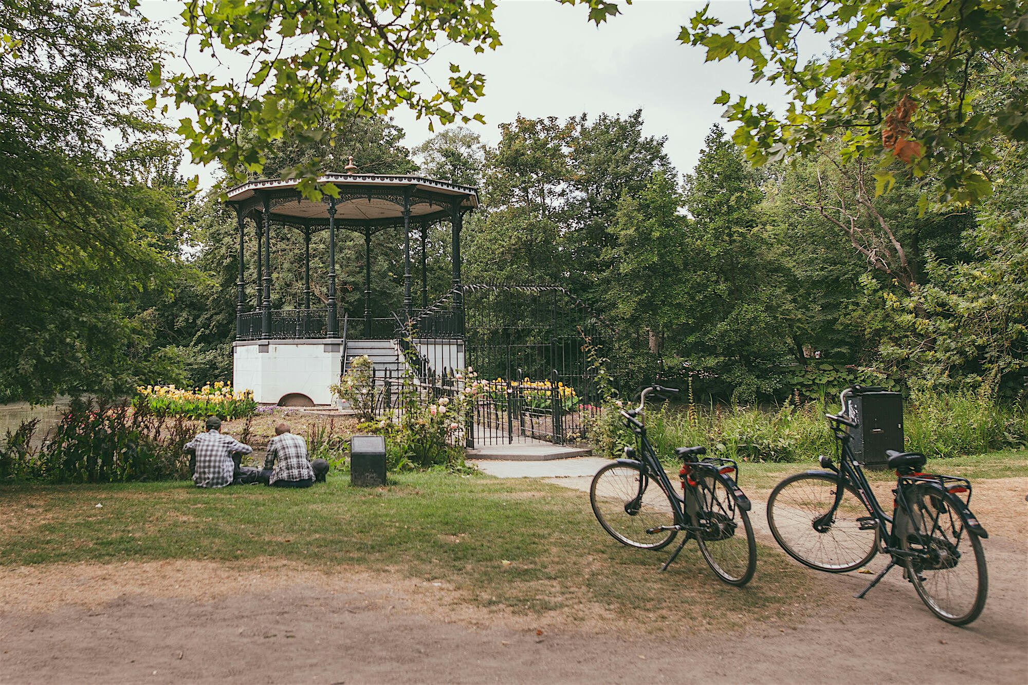Park setting with a decorative bandstand surrounded by trees and flowerbeds, with two bicycles parked on a path and people sitting on the grass
