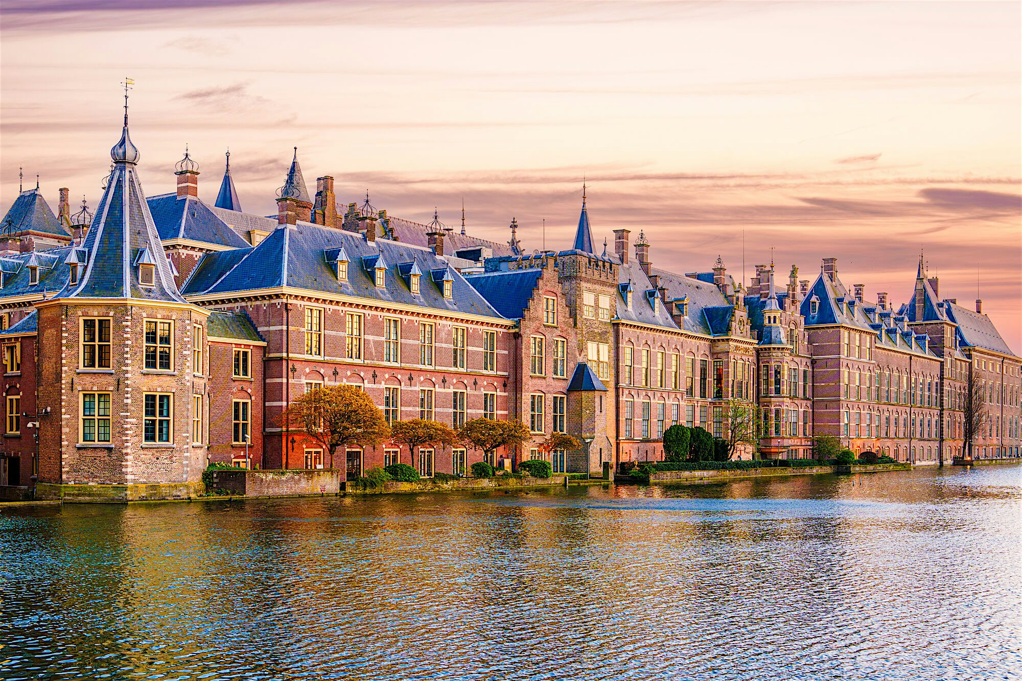 Historic waterfront complex with ornate brick buildings and blue‑roofed towers reflected in calm water,