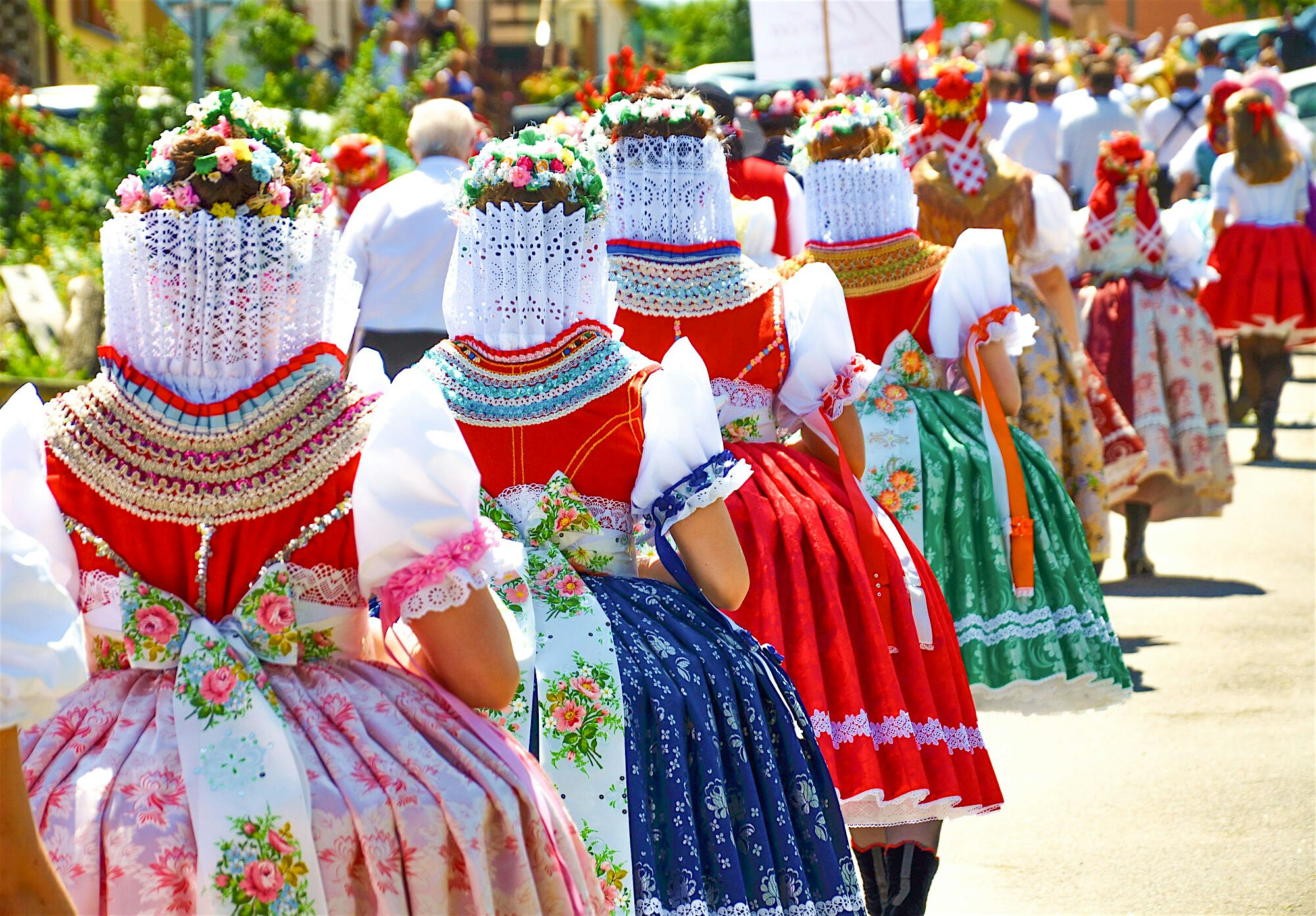 Line of people wearing brightly coloured traditional dresses and floral headpieces, walking together along a sunlit street during a parade.