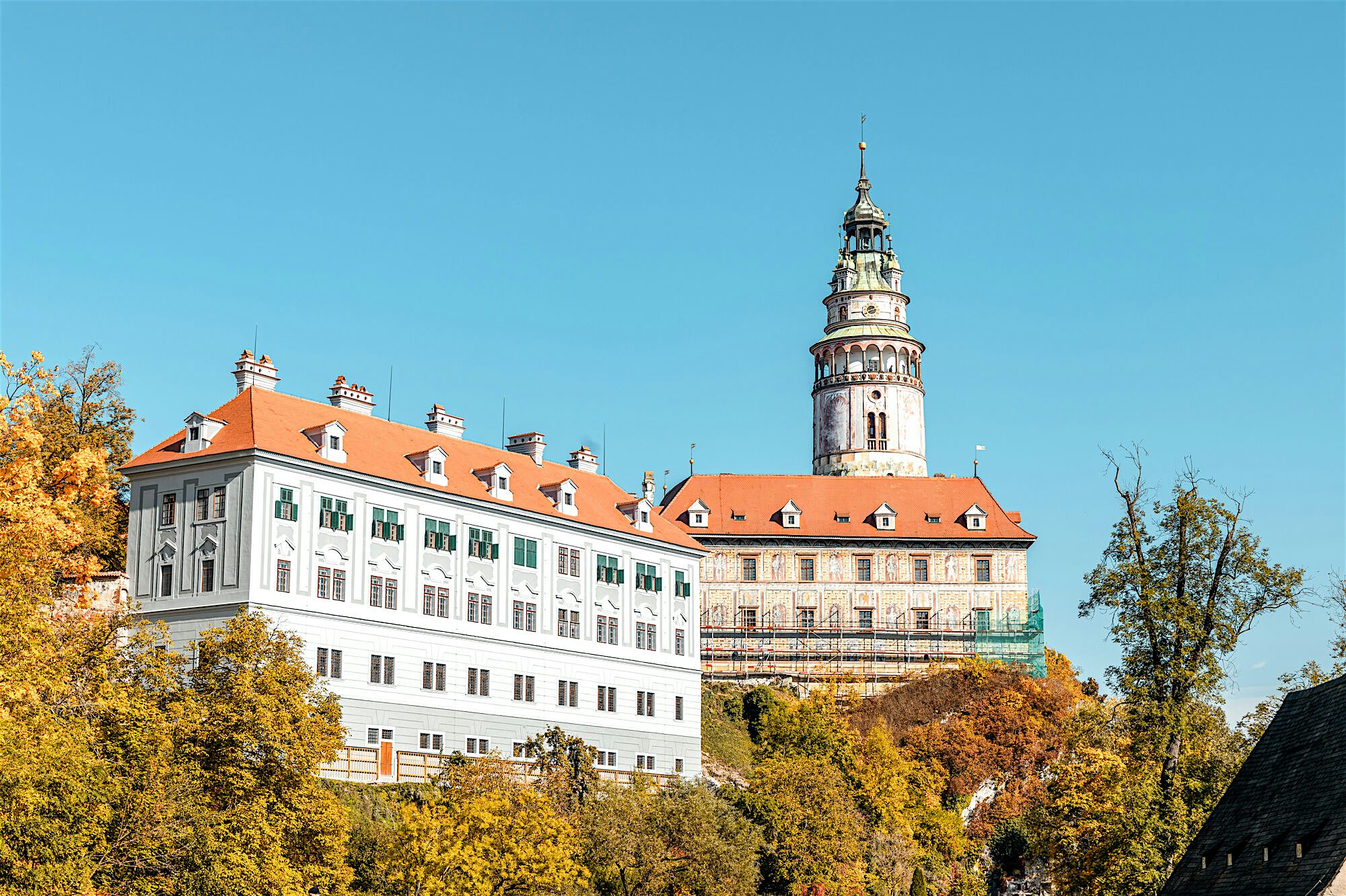 Grand hilltop castle with pale façades and red roofs rising above autumn trees against a cloudless sky.