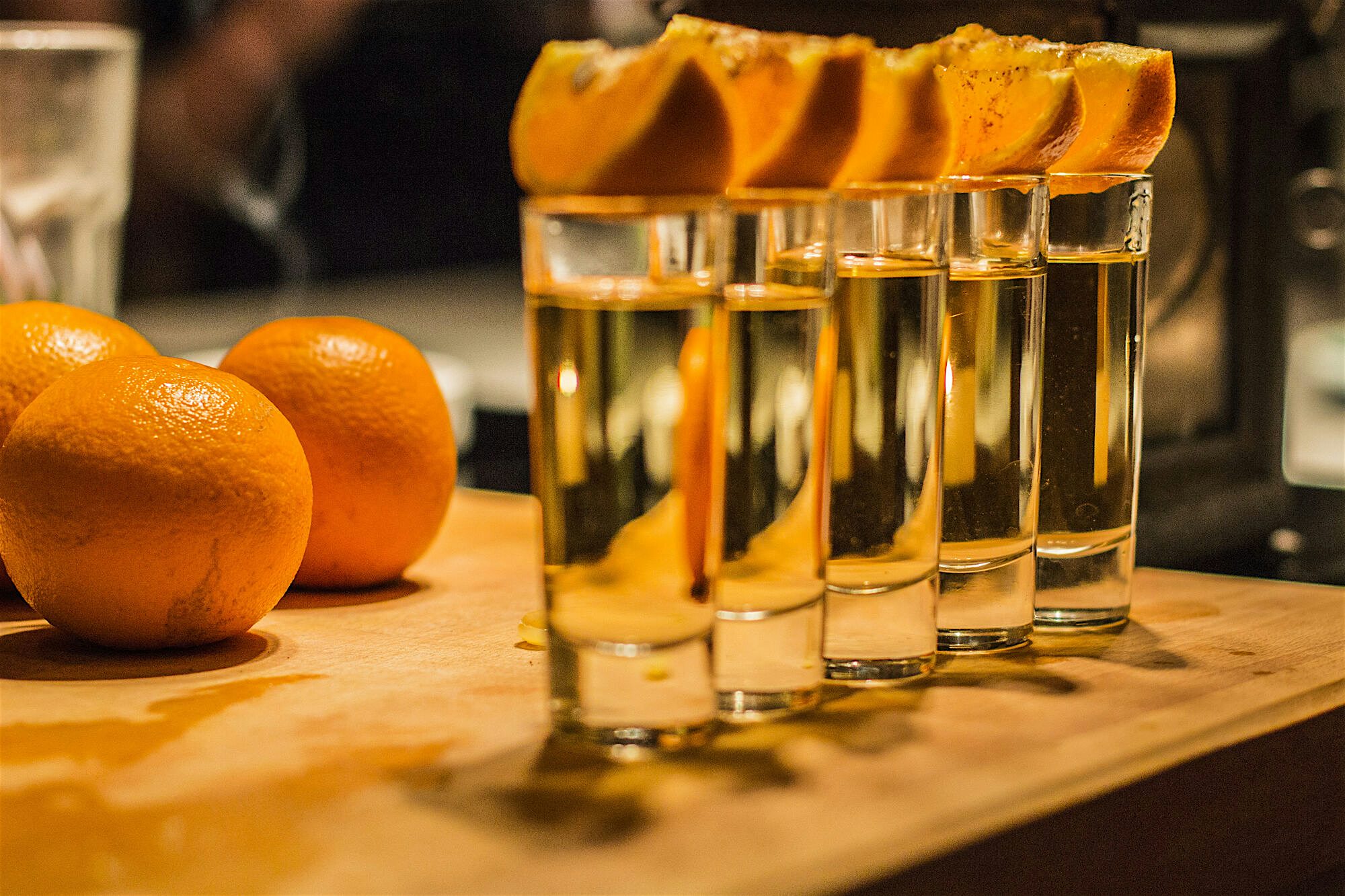 Line of clear shot glasses topped with orange slices on a wooden board, set beside fresh oranges on a softly lit bar counter.