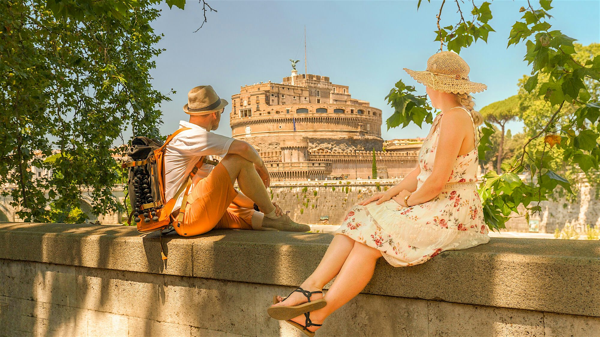 A couple sat on a wall onlooking of Castel Sant'Angelo, it is a clear day with light blue sky and a few trees sourounding the spot