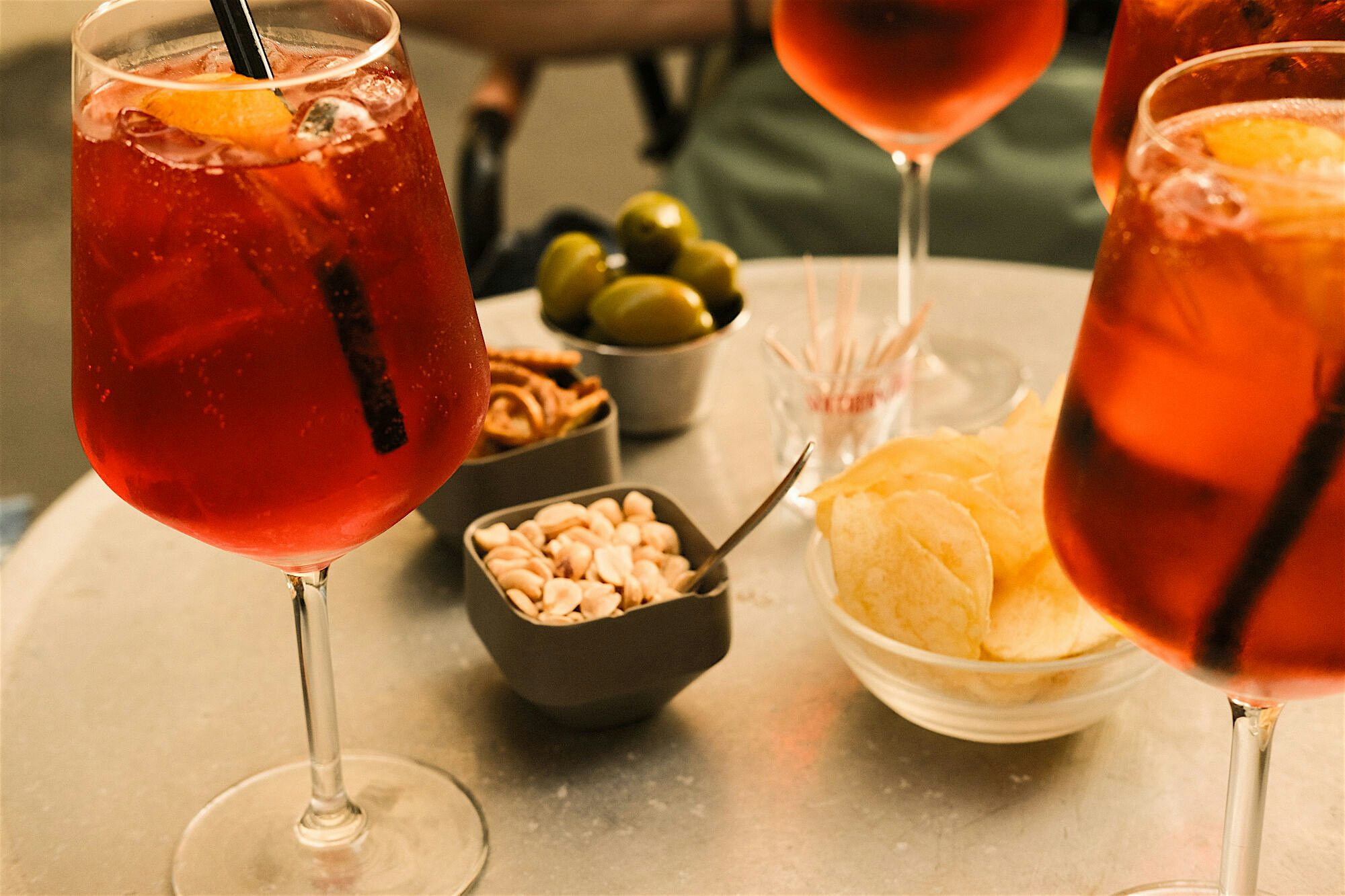 "Bright orange aperitif drinks served in glasses alongside small bowls of snacks on a café table.  "