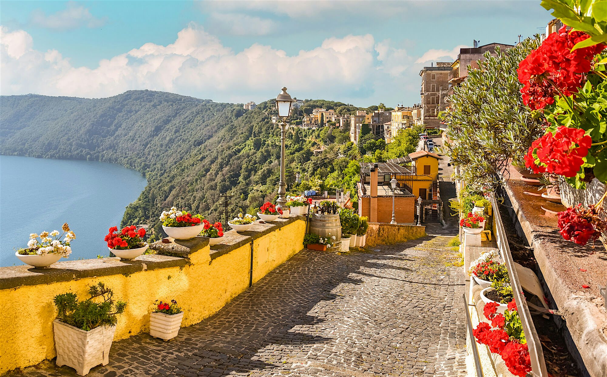 "A cobbled path lined with colourful flowers overlooking a lake, with hills and buildings in the background.  "