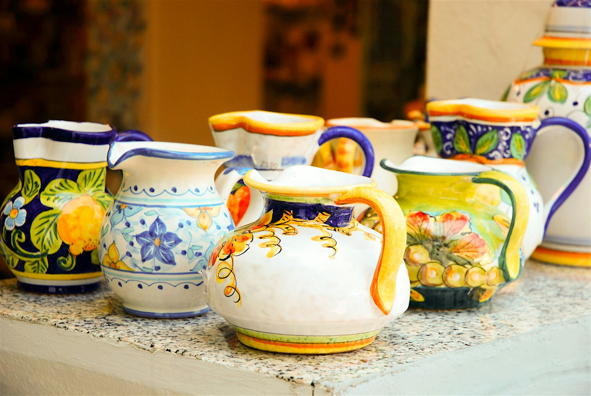 Collection of handmade and handpainted jugs. They are painted in yellow and shades of blue, with traditional designs featuring flowers, and fruit such as lemons.