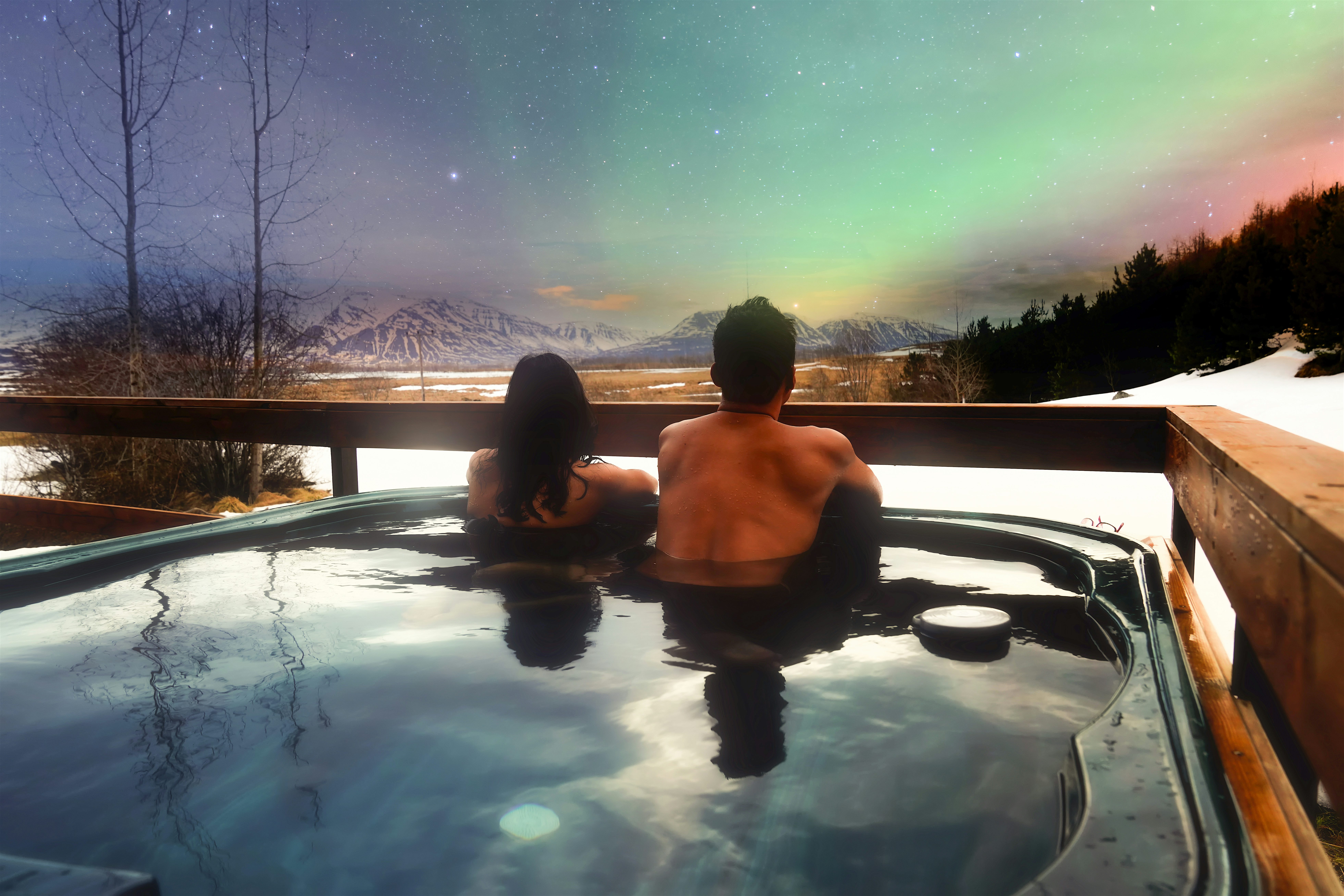 Couple outside in a private hot tub viewing the colourful Northern Lights above with snow in front of them and trees to the sides.