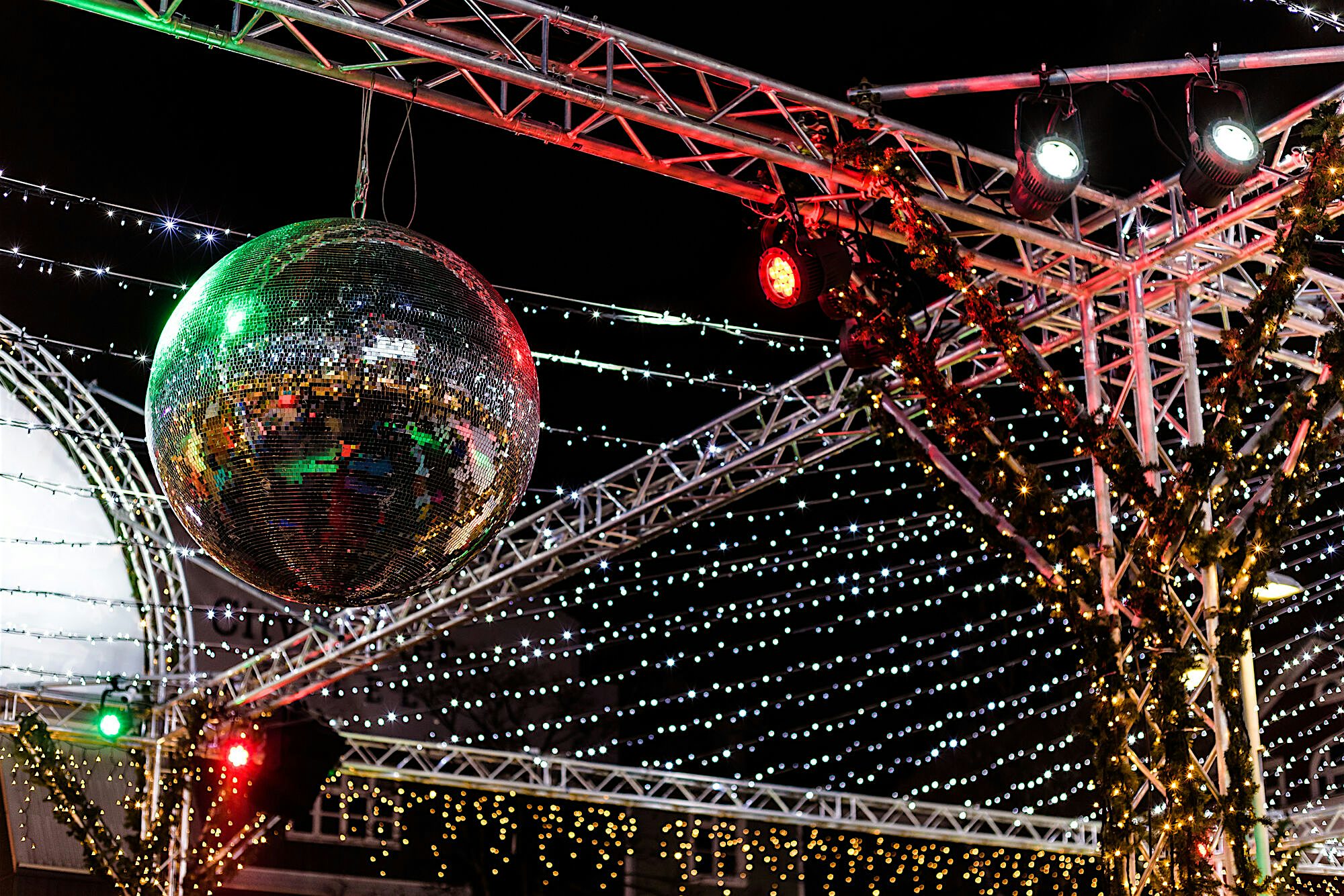 Large disco mirror ball hanging from metal staging with strings of lots of small yellow and white lights