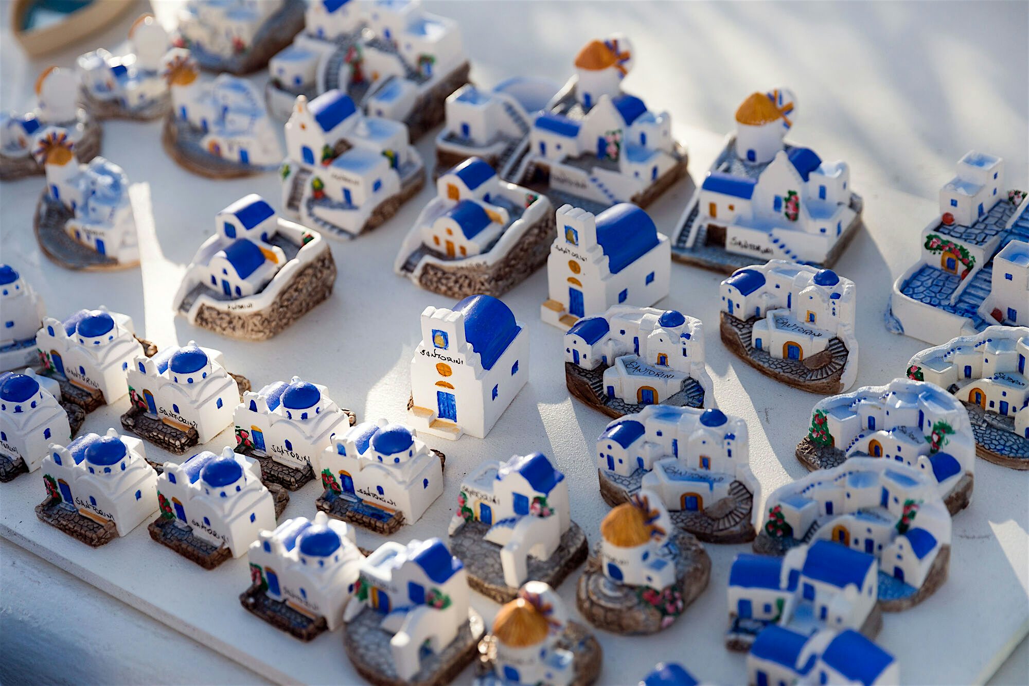 Display of small ceramic souvenir models shaped like white houses with blue domes and roofs, arranged closely together on a table to resemble a hillside village.