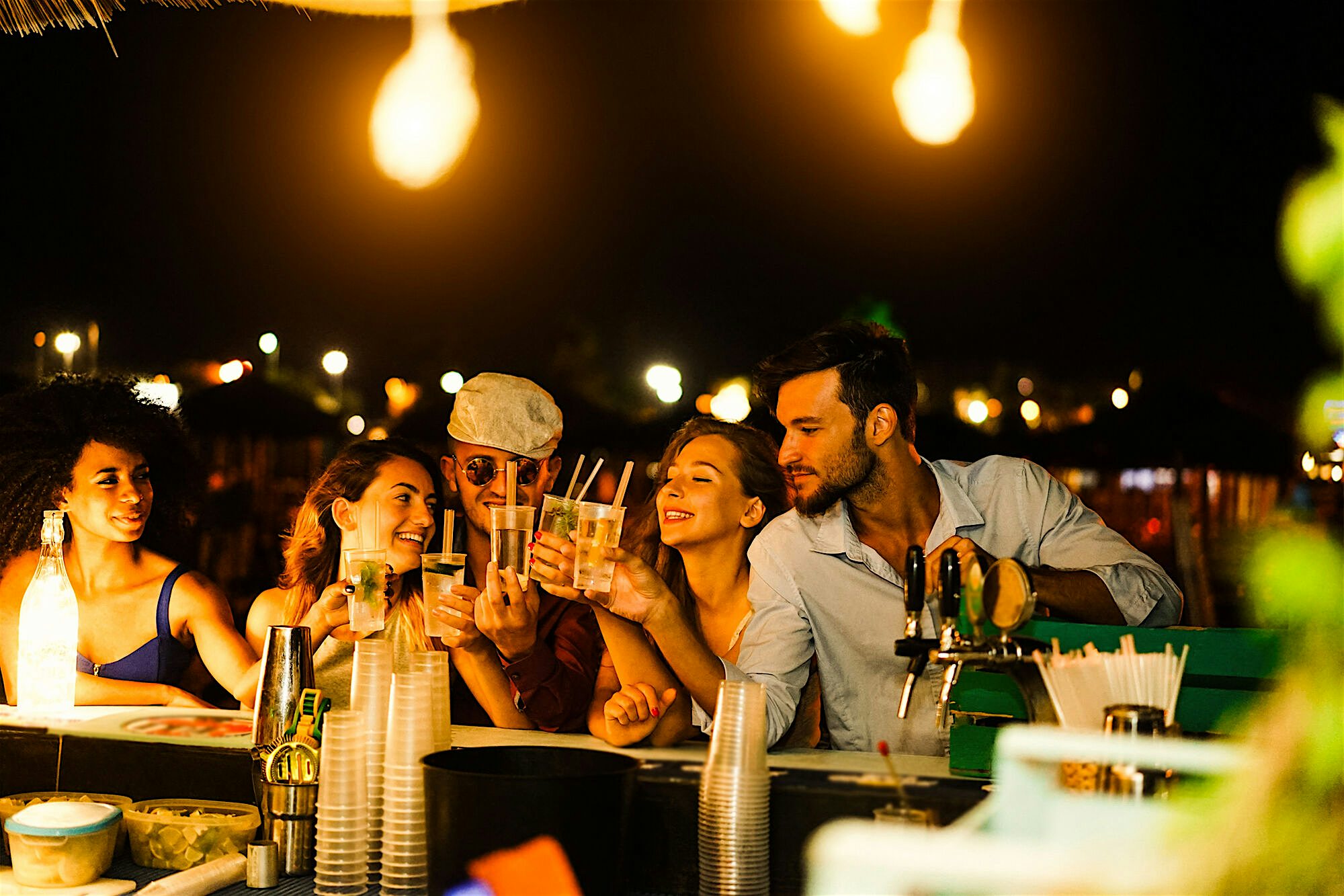 Group of people gathered at an outdoor bar at night, holding drinks across the counter, with glassware, bottles, and bar equipment