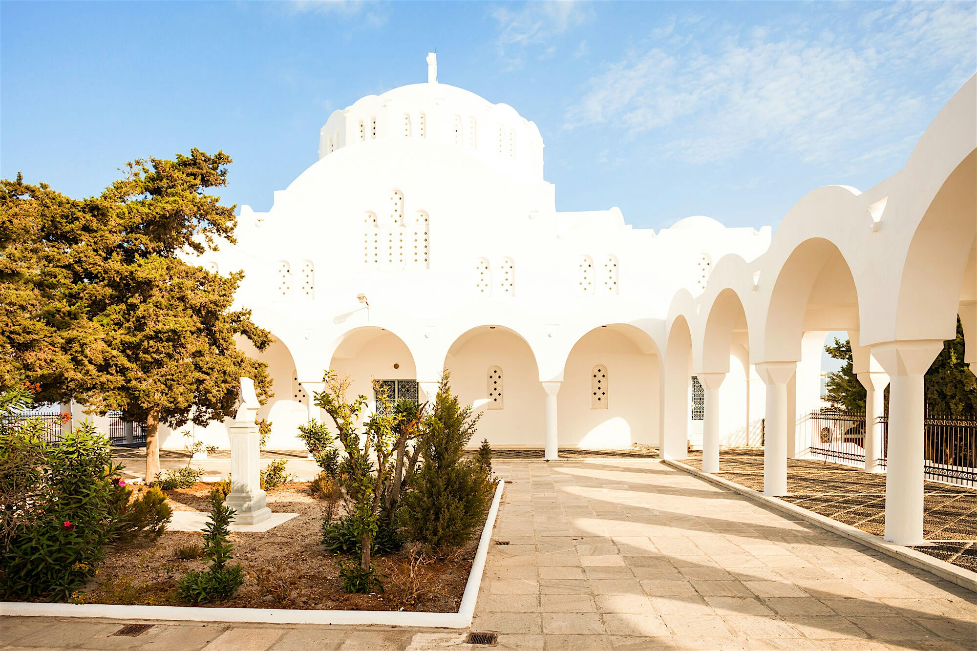 Whitewashed church courtyard with sweeping arched colonnades and a domed roof, featuring sunlit stone paving, small landscaped areas with shrubs and trees
