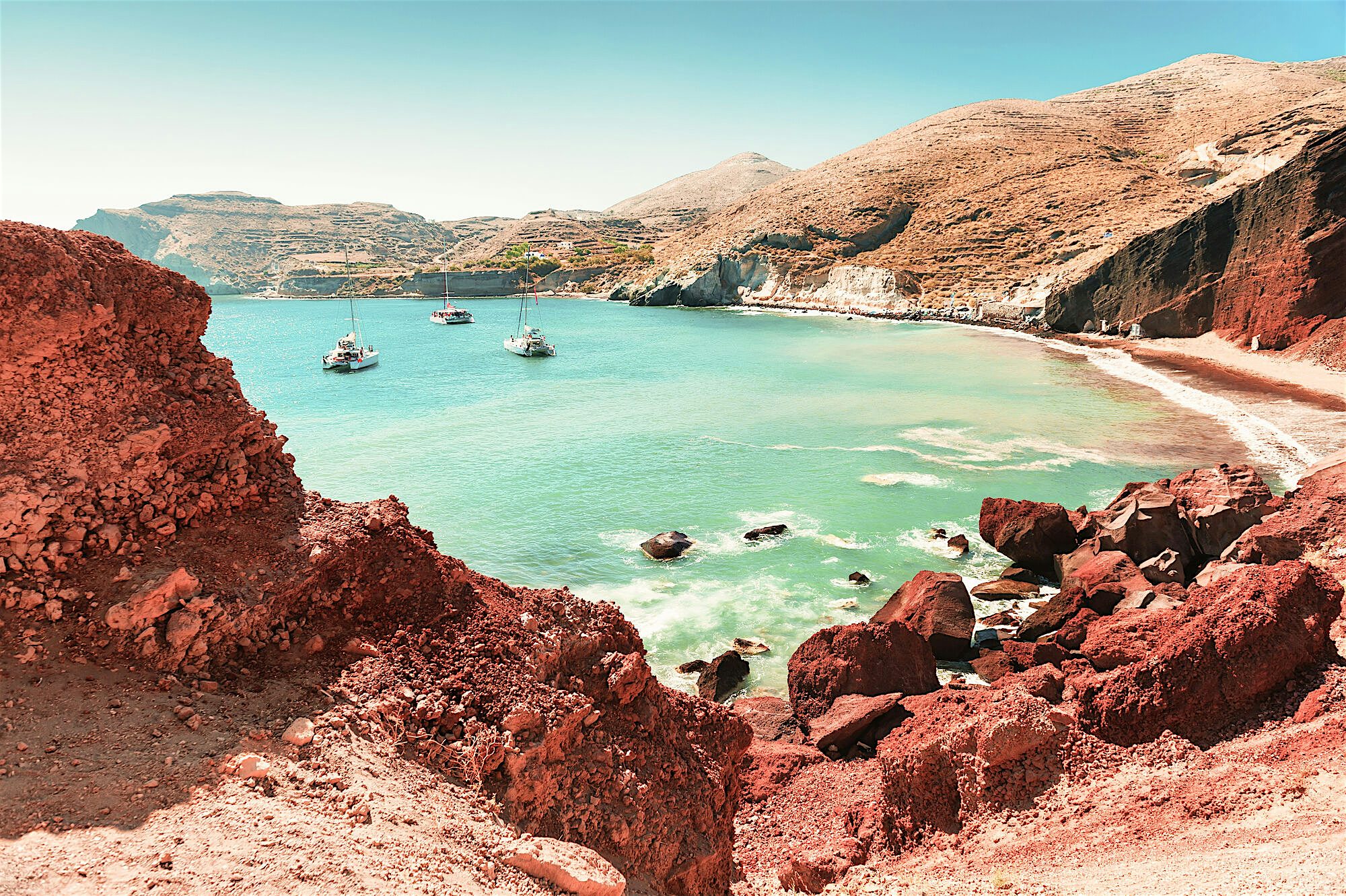 Rocky coastal cove with red cliffs surrounding clear turquoise water, with several small boats anchored offshore