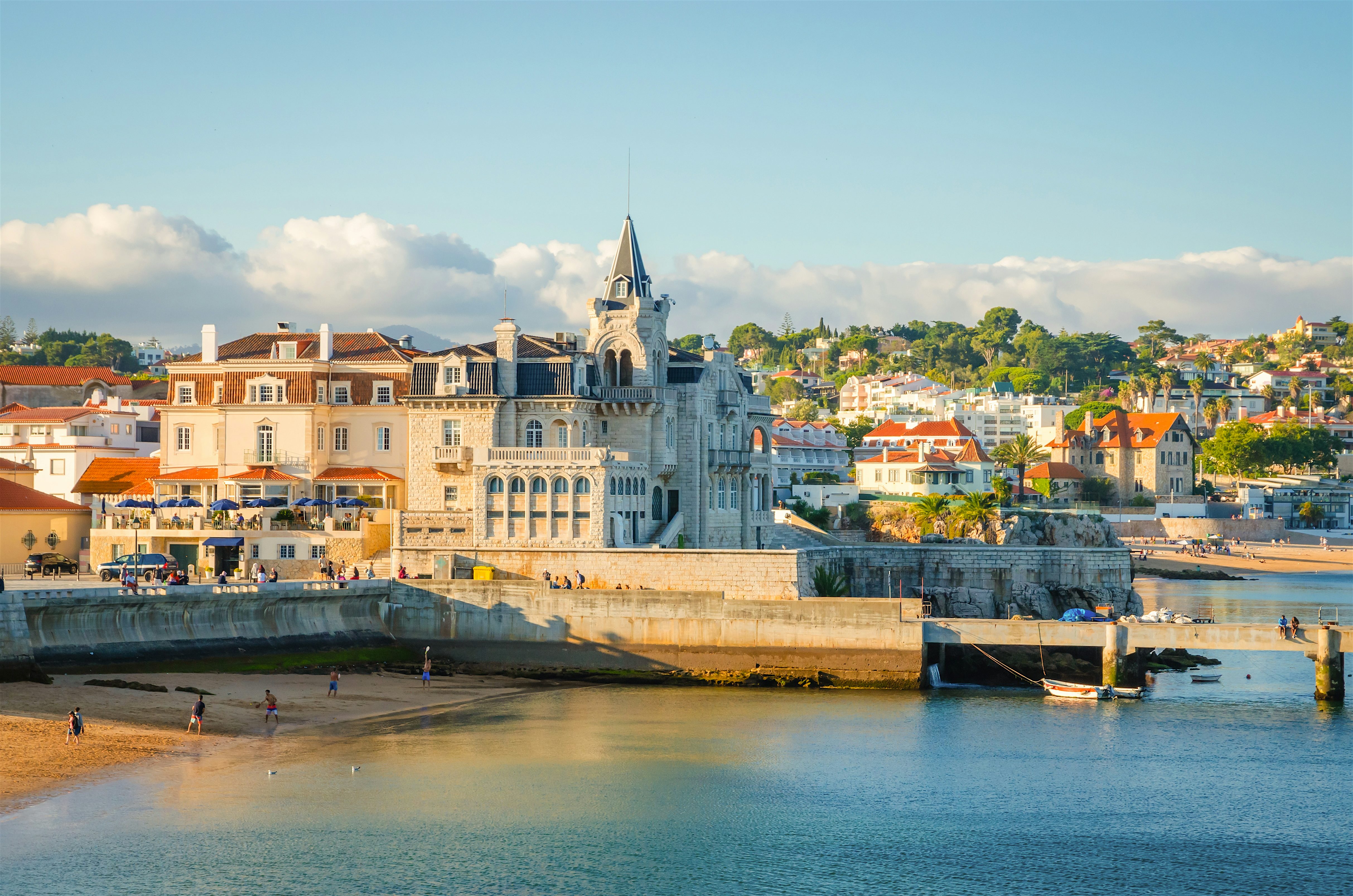 Coastal town with a grand seaside building and sandy beach, backed by pastel houses and greenery along a calm shoreline.