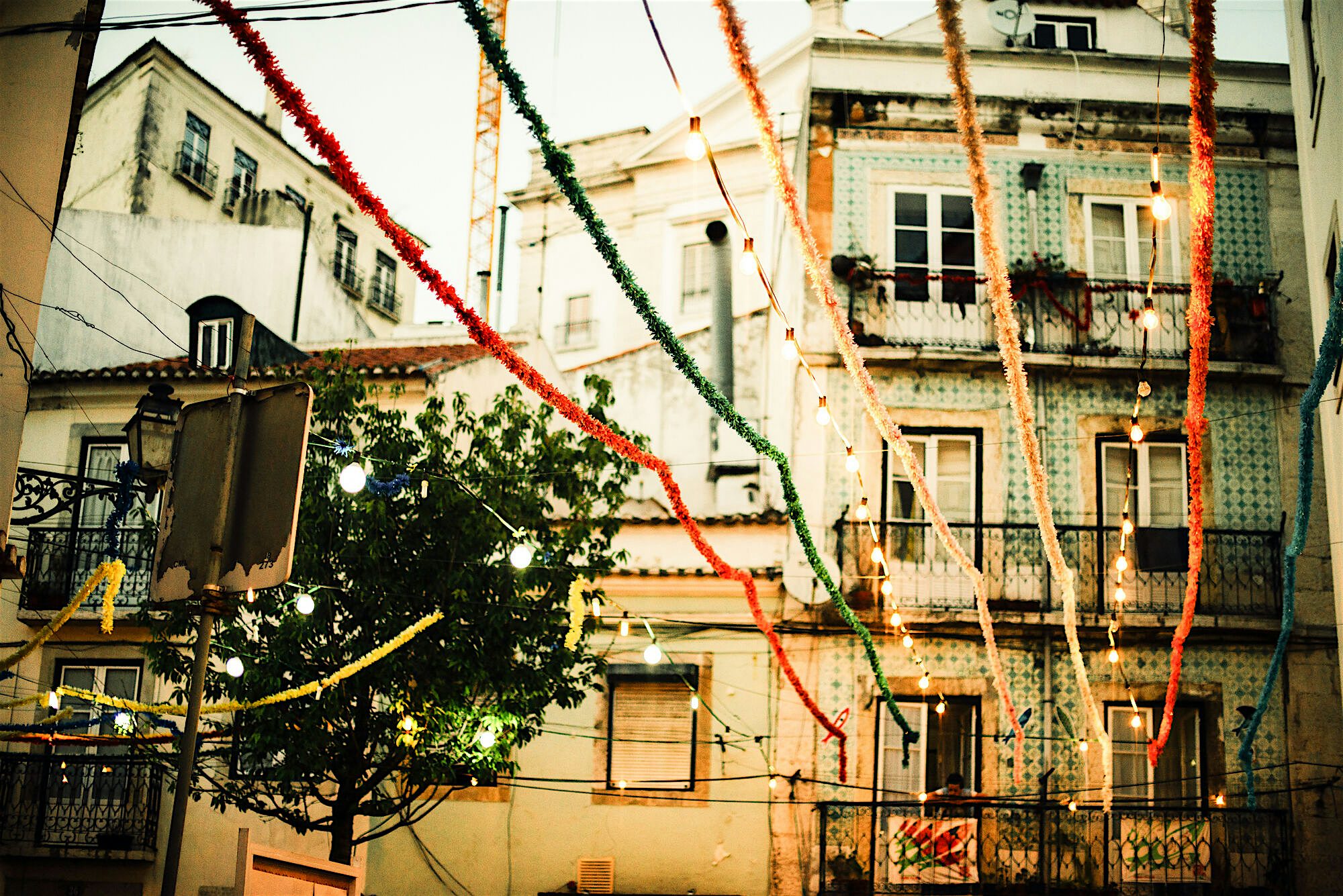 Narrow street decorated with colourful hanging streamers and lights, draped between old tiled buildings in a lively neighbourhood.