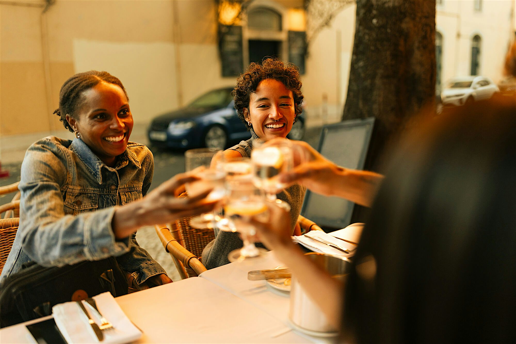 People seated at an outdoor restaurant table clinking glasses over a neatly set meal on a quiet city street.