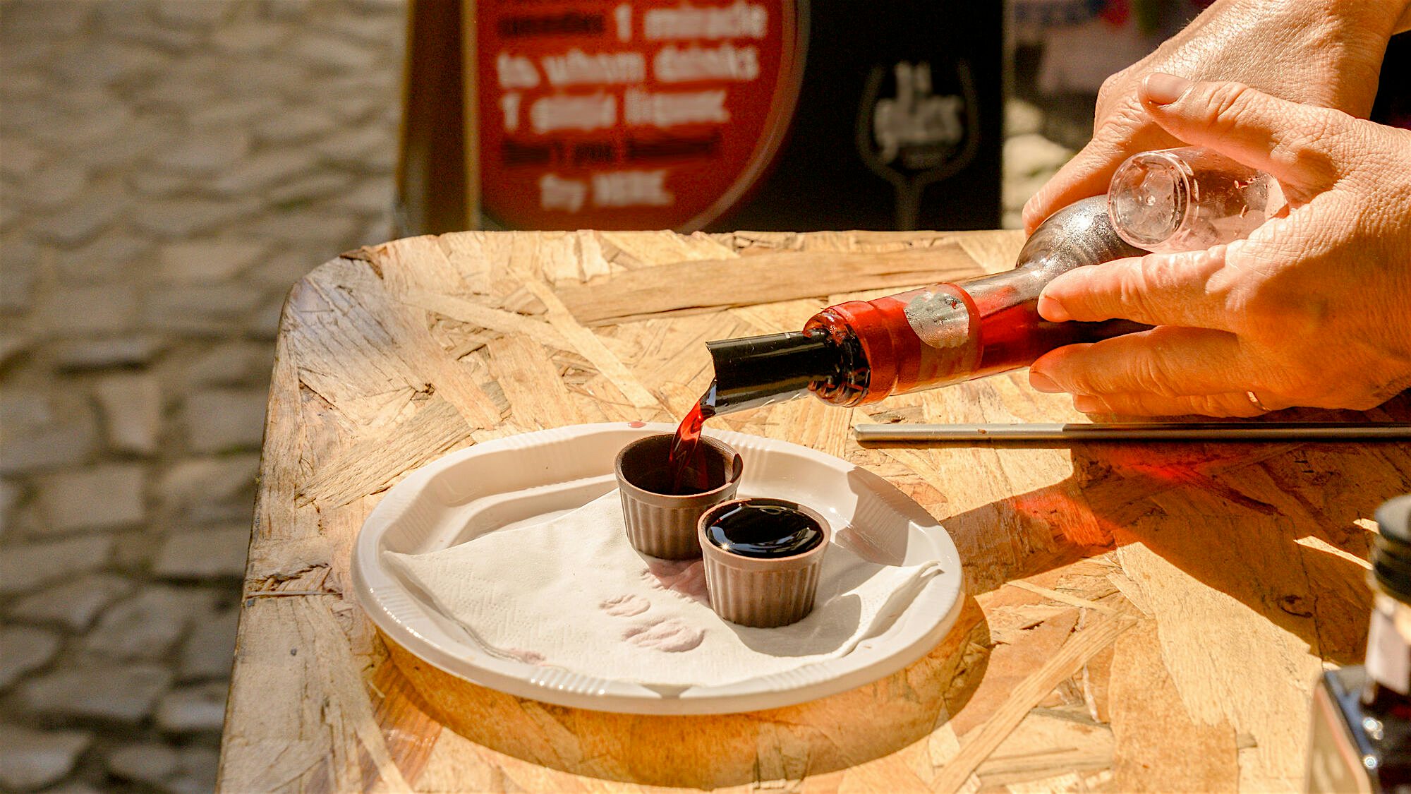 A small plate set on a wooden barrel as red liqueur is poured into tasting cups outdoors in bright sunlight.