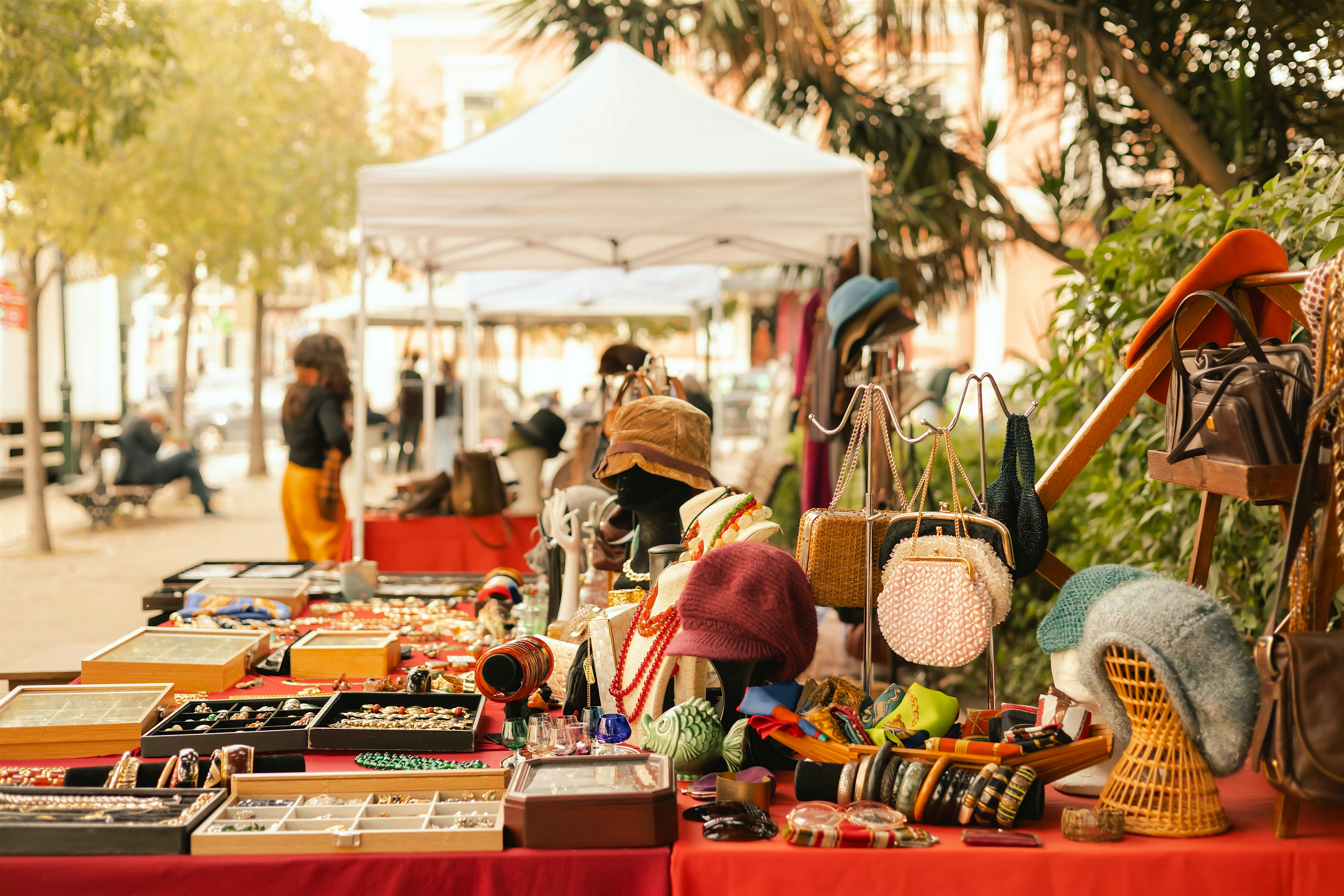 Outdoor market stall displaying jewellery, hats, handbags, and knitted accessories laid out on red tablecloths beneath a white canopy.