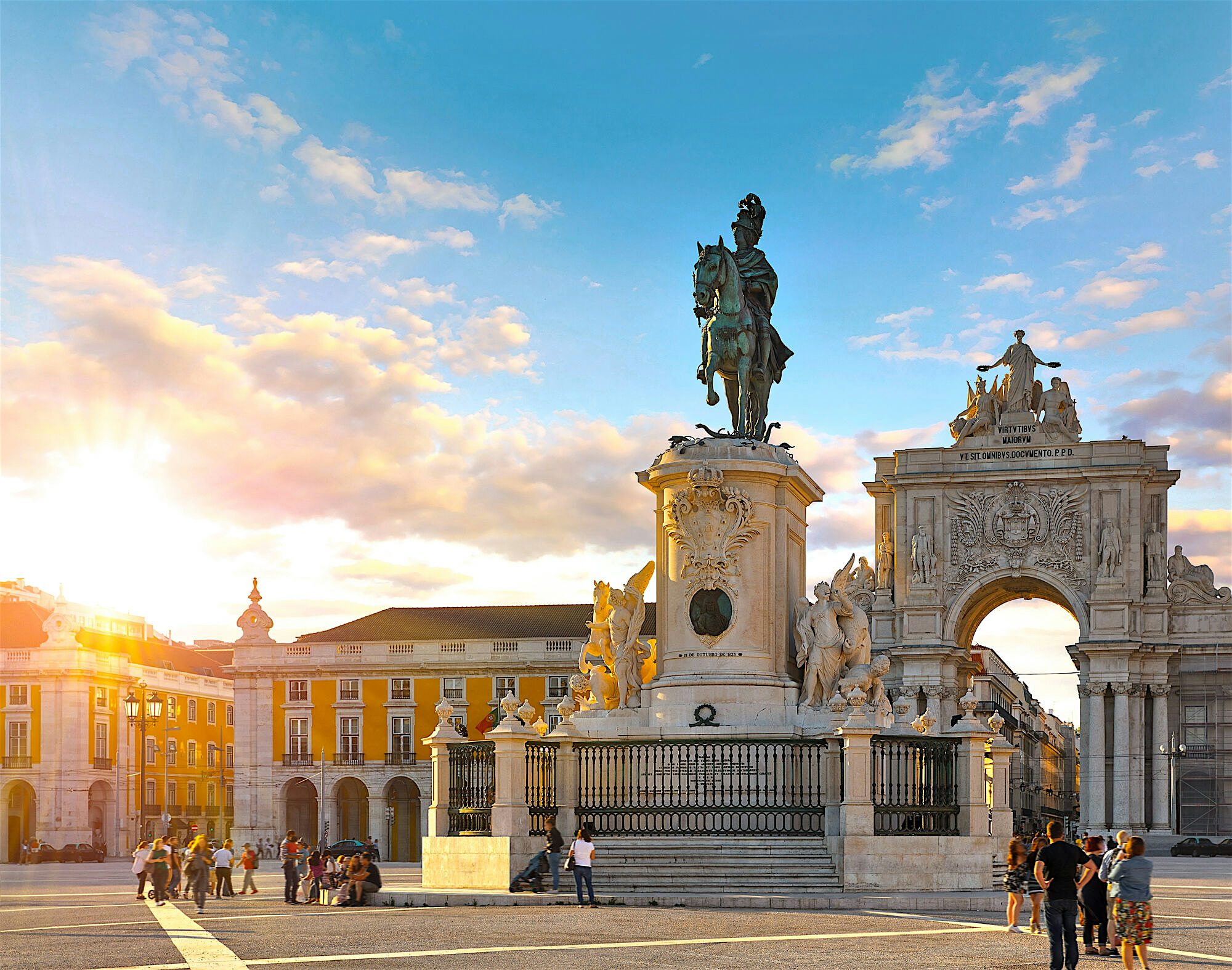 Large stone square with an equestrian statue at its centre, surrounded by historic buildings and an ornate triumphal arch glowing in warm evening light.