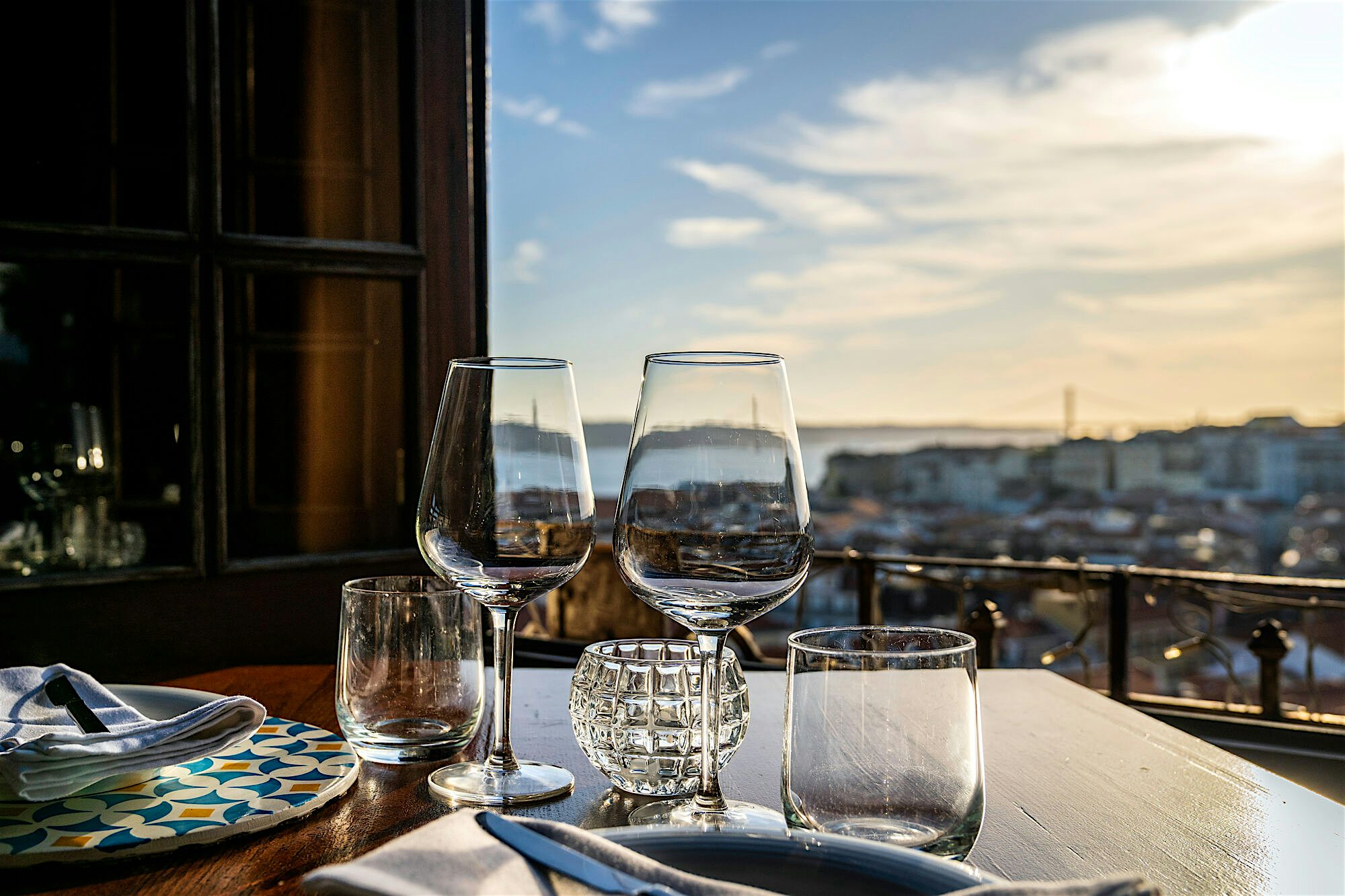 Table set with clear wine glasses and cutlery on a wooden surface, overlooking a city skyline at sunset from a balcony or terrace.