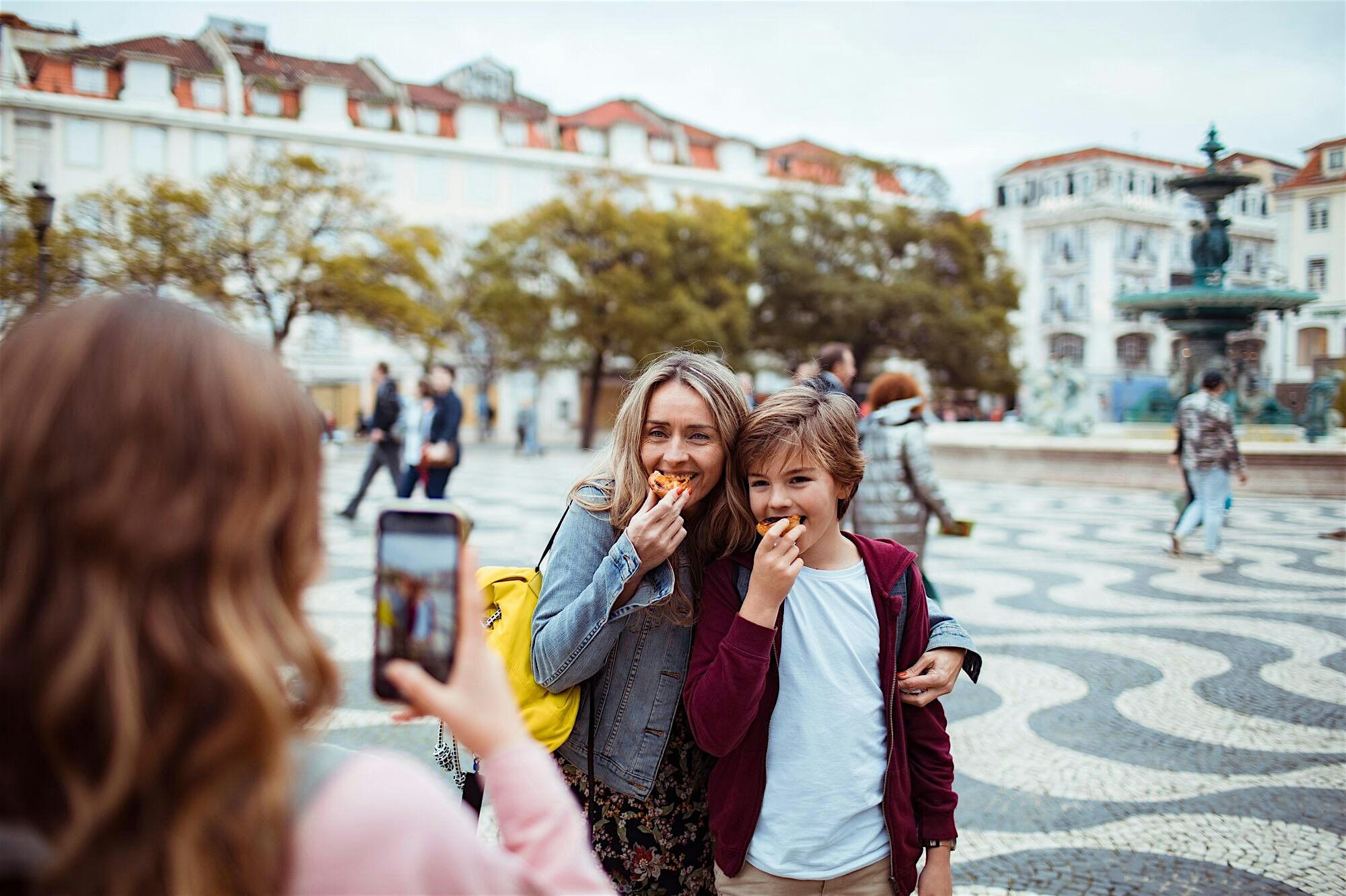 Two people posing while eating pastries in a large tiled square, with a fountain and city buildings in the background as someone takes a photo.