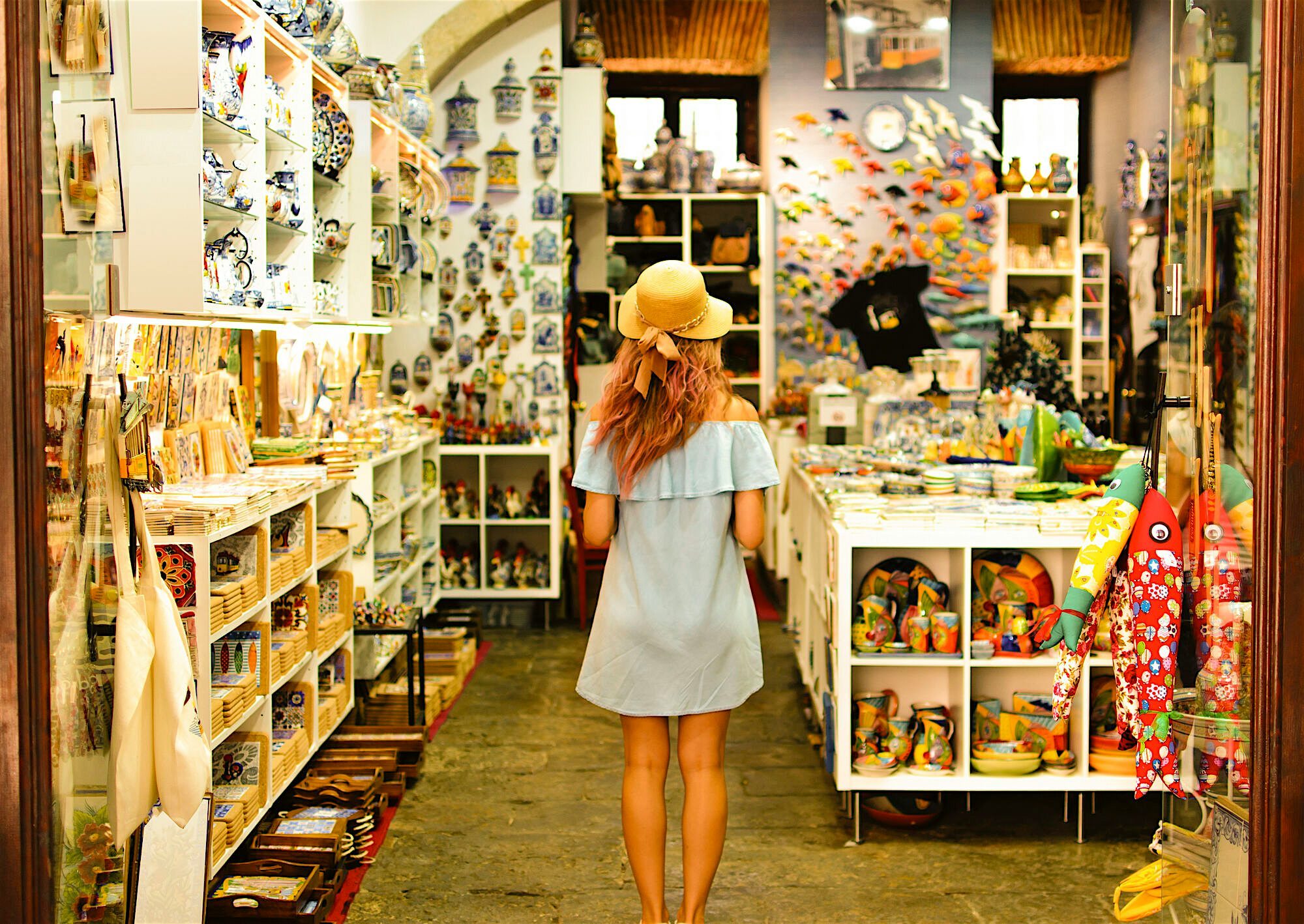 Person standing inside a small souvenir shop filled with ceramics, magnets, textiles, and colourful handmade items, browsing shelves arranged tightly along a narrow aisle.