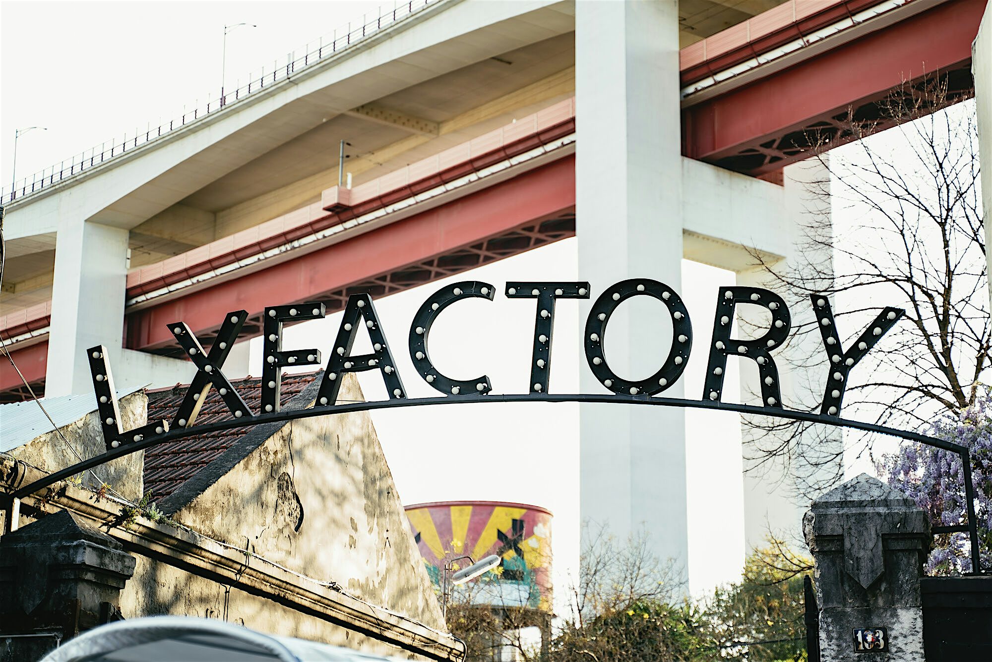 Metal gateway sign reading “LX Factory” beneath an elevated bridge, set against industrial structures and street art.