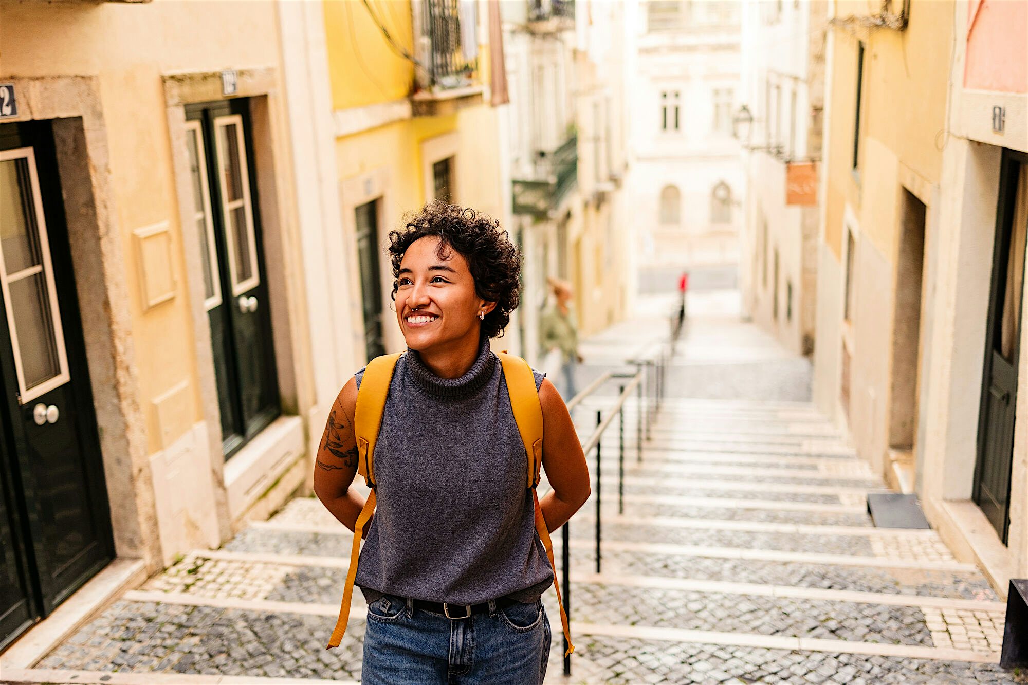 Person standing on stone steps between pastel-coloured buildings, wearing a backpack and looking out over a descending residential street.